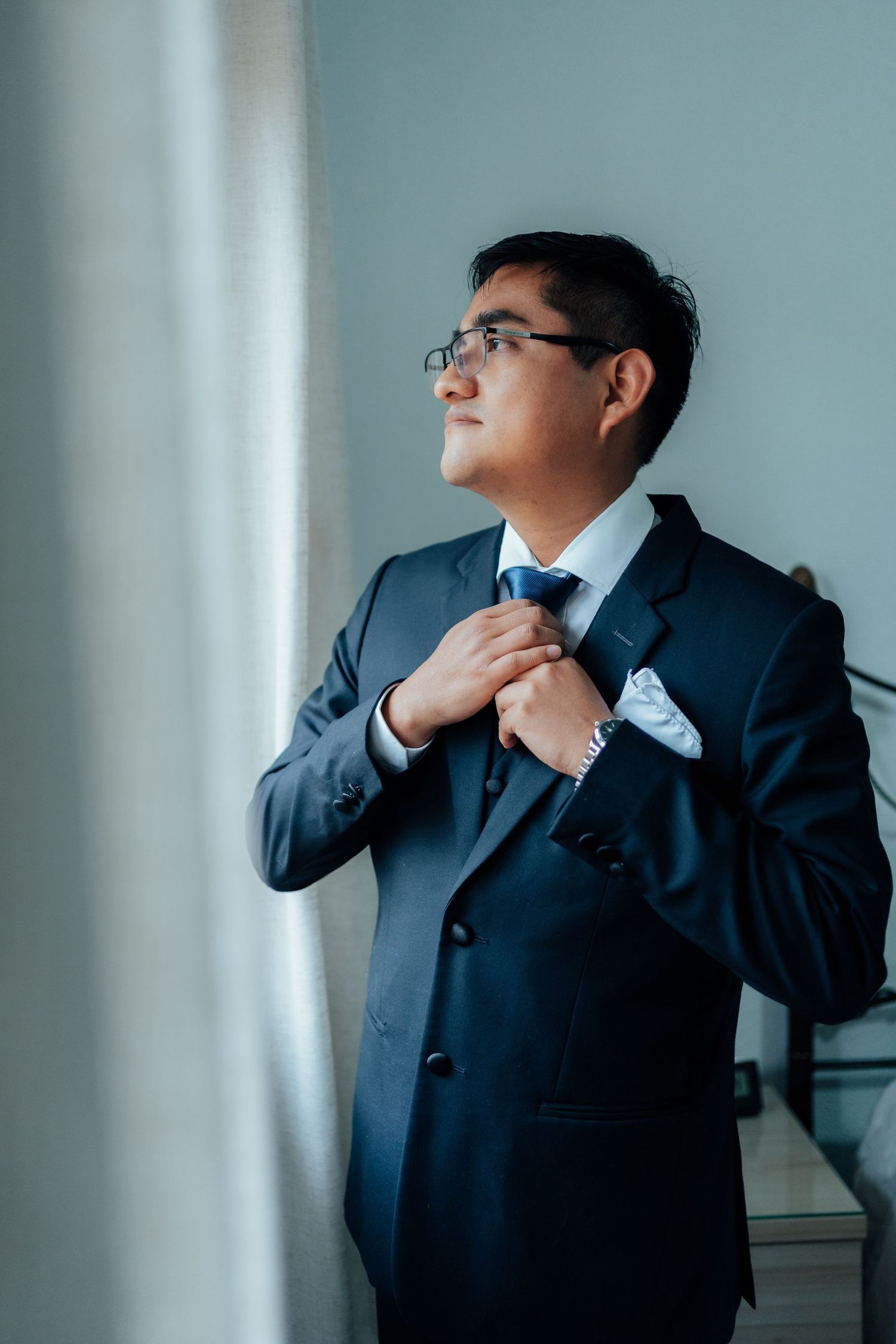 A well-dressed groom in a navy suit adjusts his tie while gazing out a window.