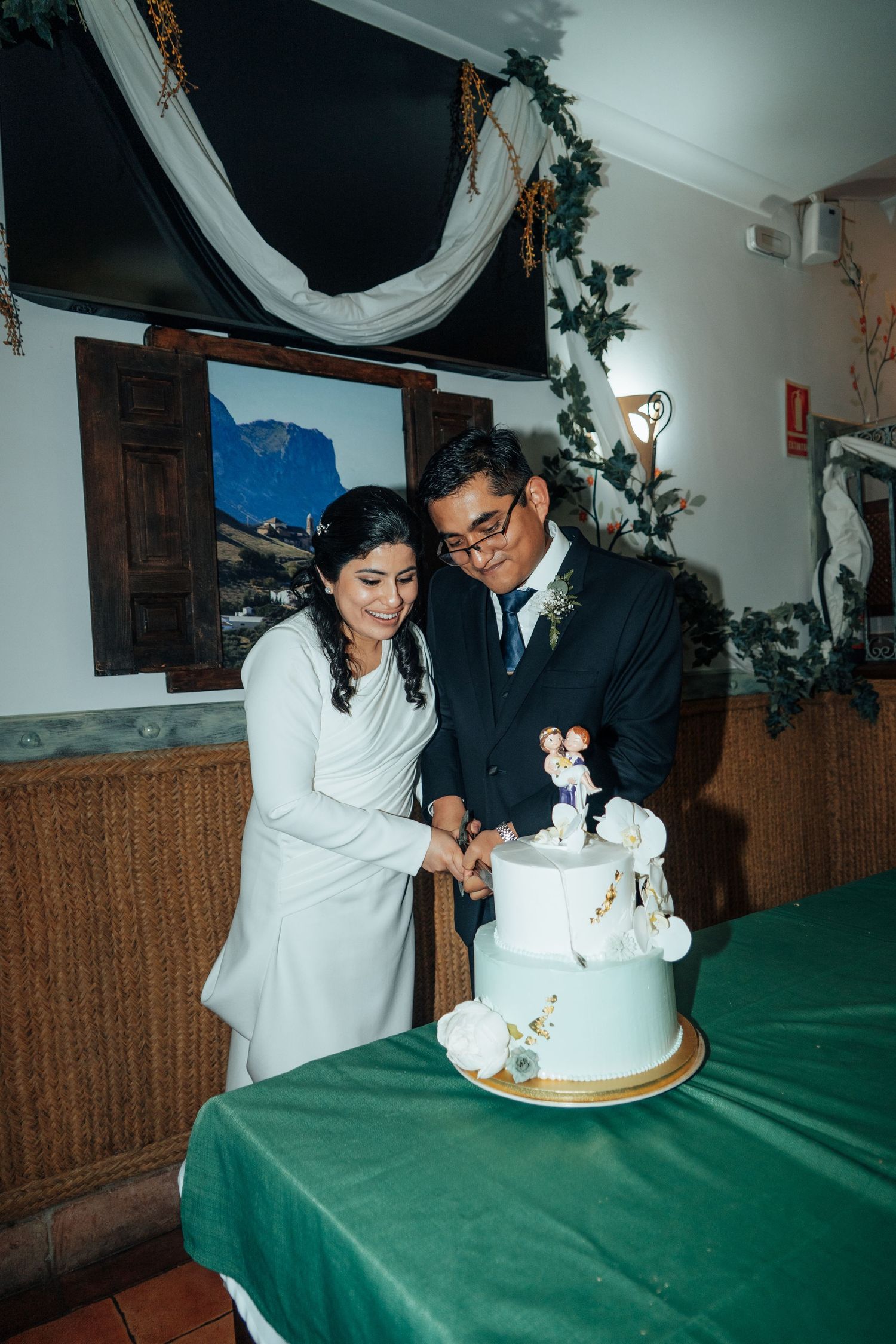 Bride and groom cutting a two-tier white wedding cake decorated with floral toppers together.