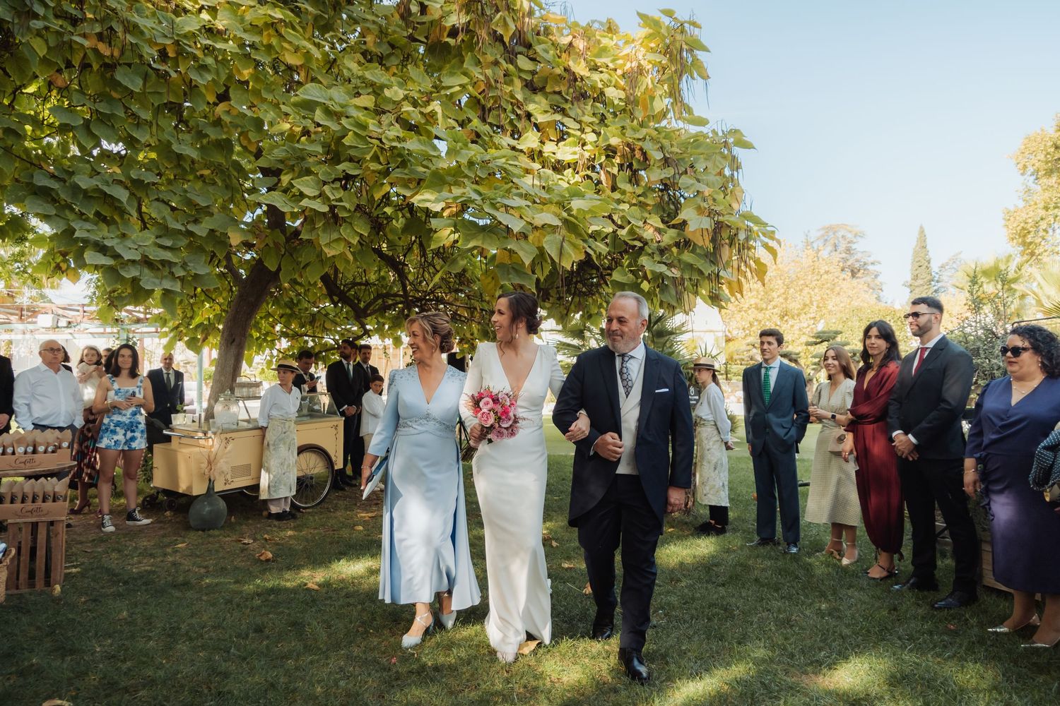 Bride in white gown walks aisle with parents past smiling guests at garden wedding.