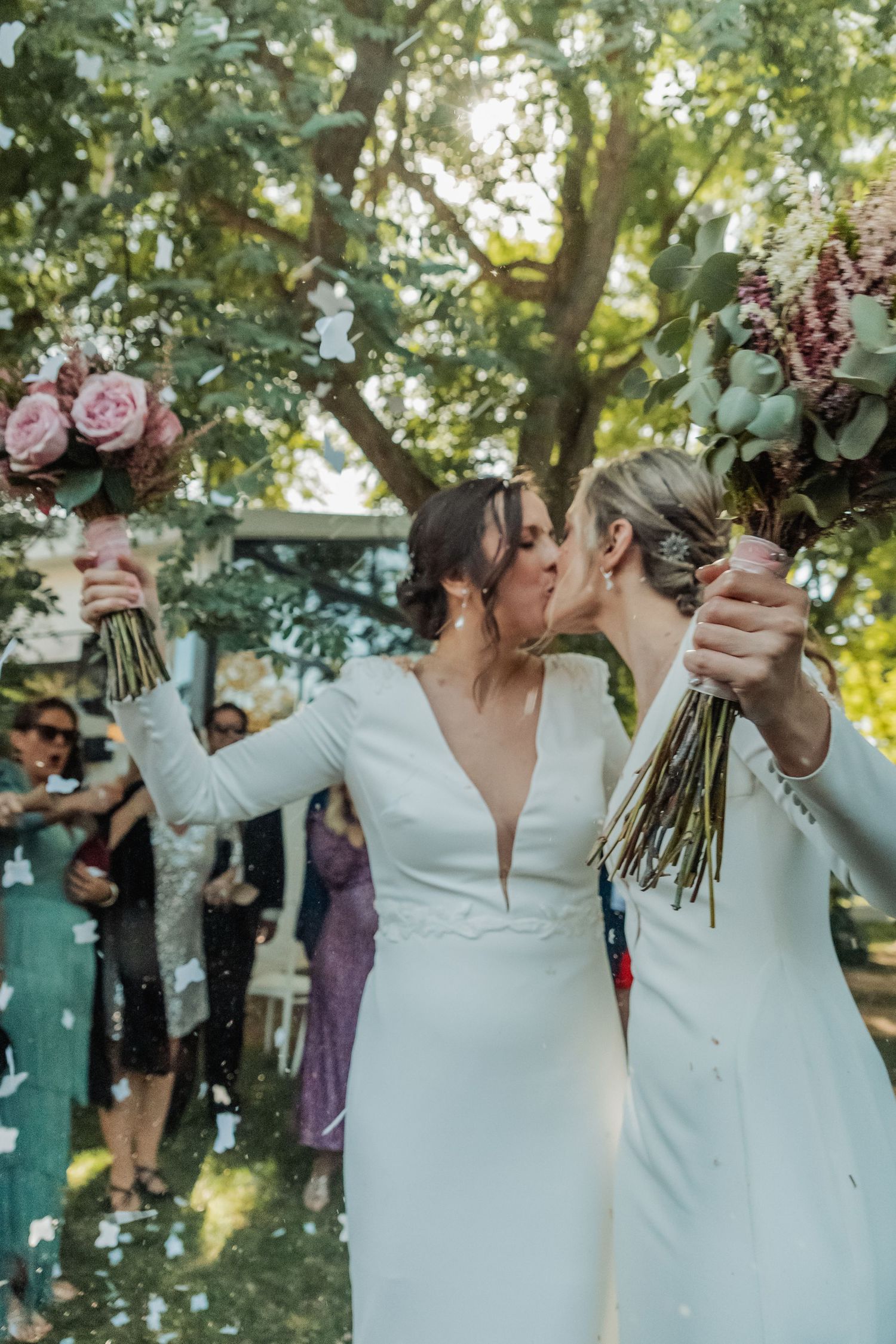 Two brides in white gowns kiss and raise bouquets while guests celebrate with confetti at outdoor wedding.