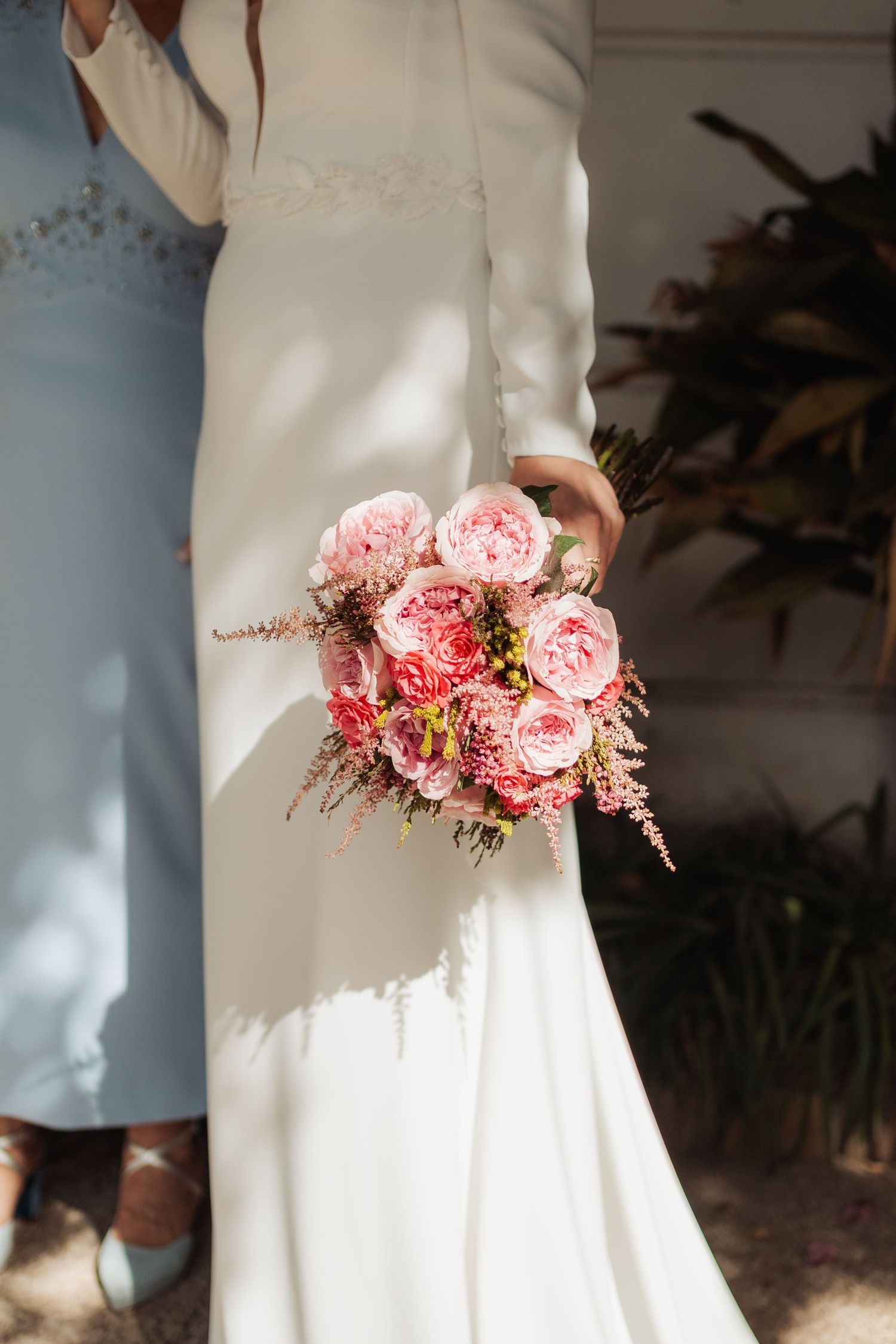 Close-up of bride holding a lush pink peony and rose wedding bouquet against white wedding dress outdoors.