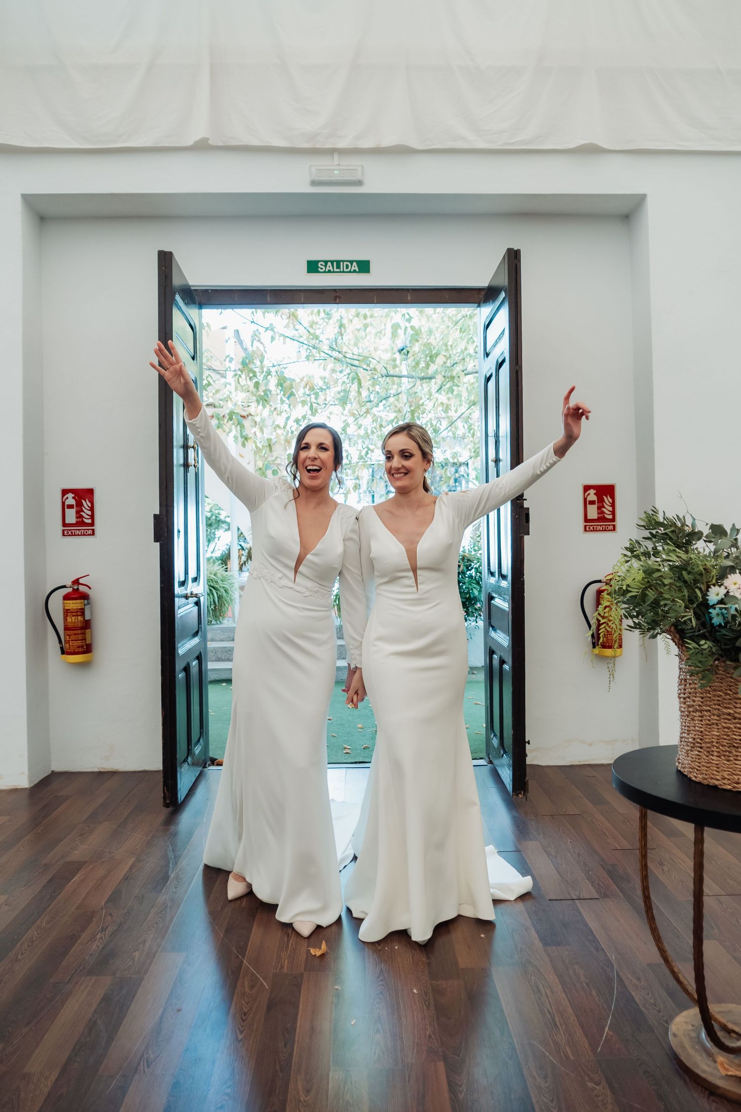 Two brides in elegant white long-sleeve wedding gowns celebrating at venue entrance with arms raised.