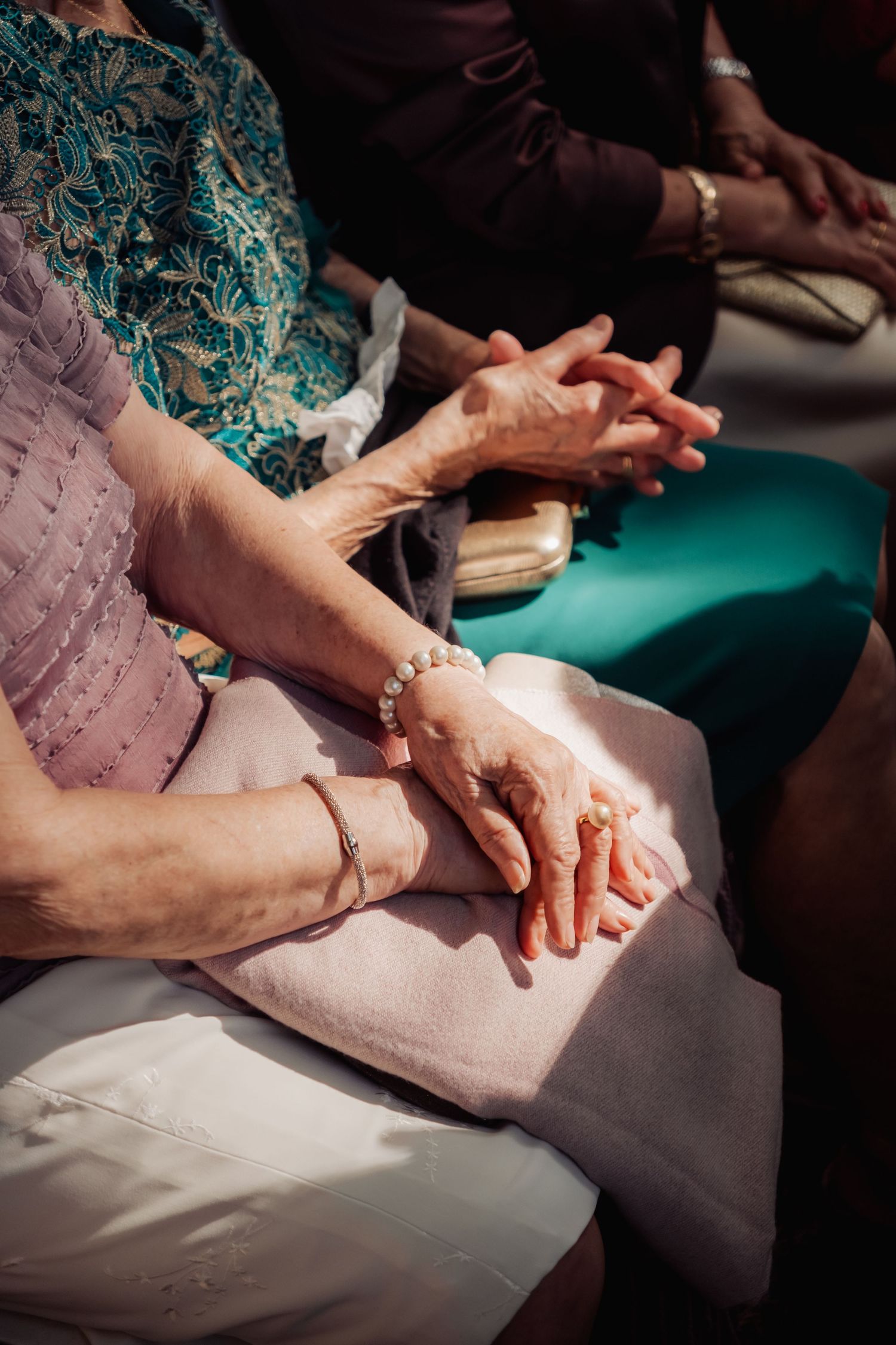 Close-up of elderly women's hands with bracelets resting on laps during a wedding ceremony.
