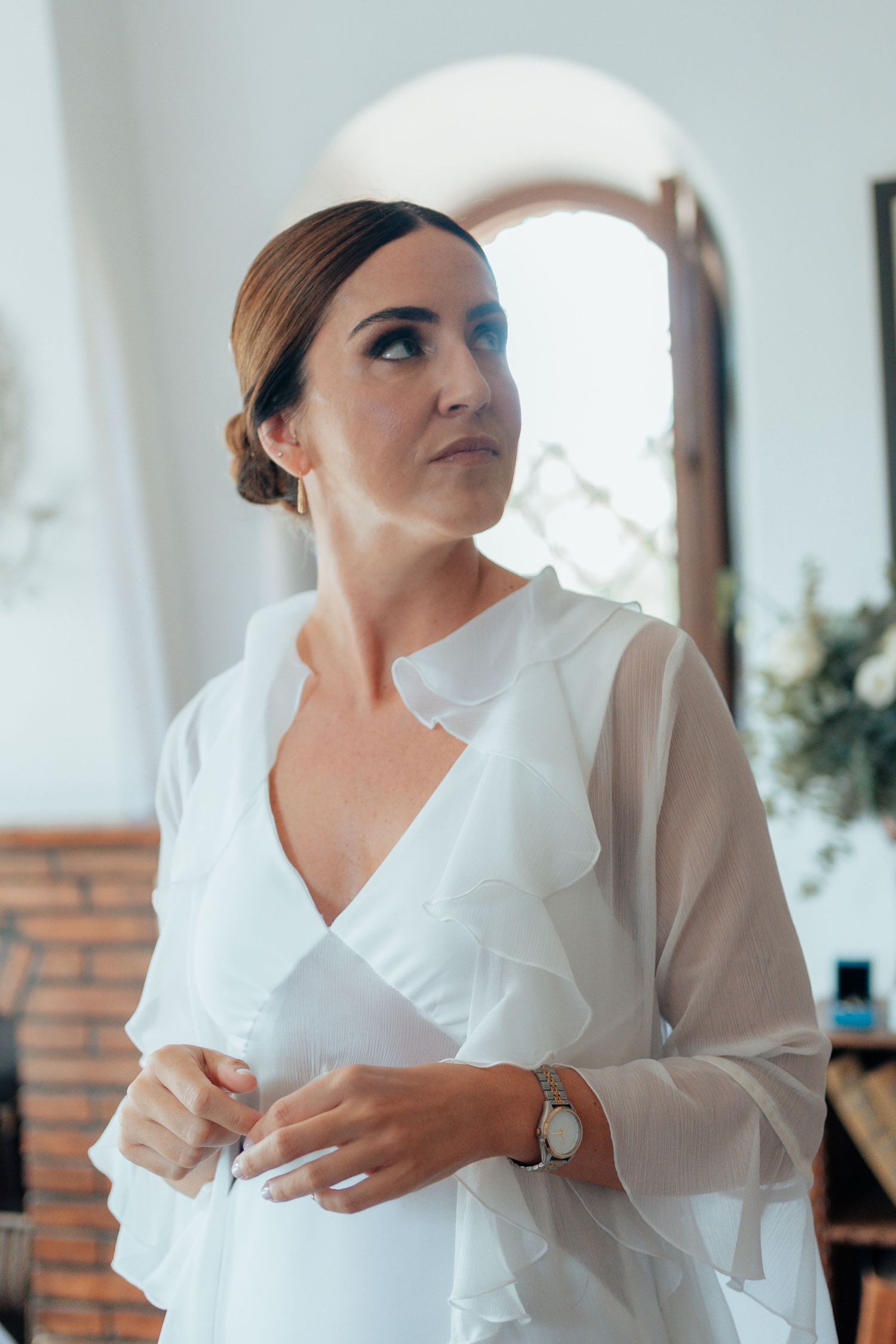 Elegant bride in white chiffon robe adjusting her outfit before wedding ceremony indoors.