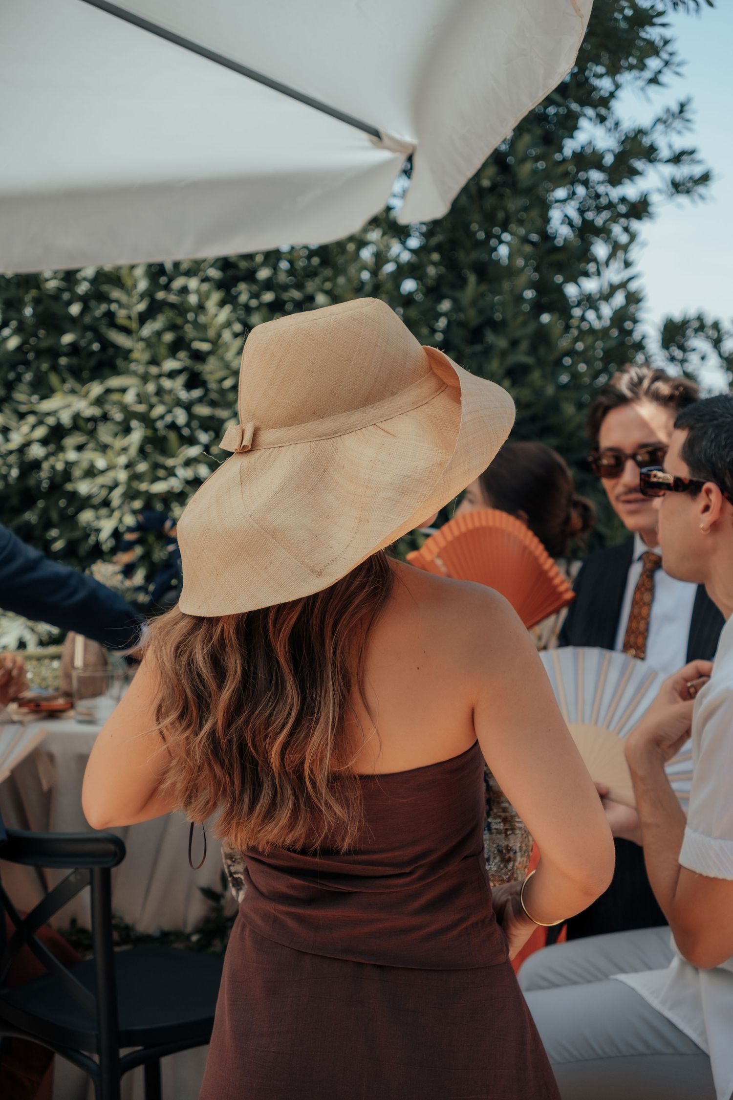 Woman in brown strapless dress and wide-brim straw hat at outdoor summer wedding reception.