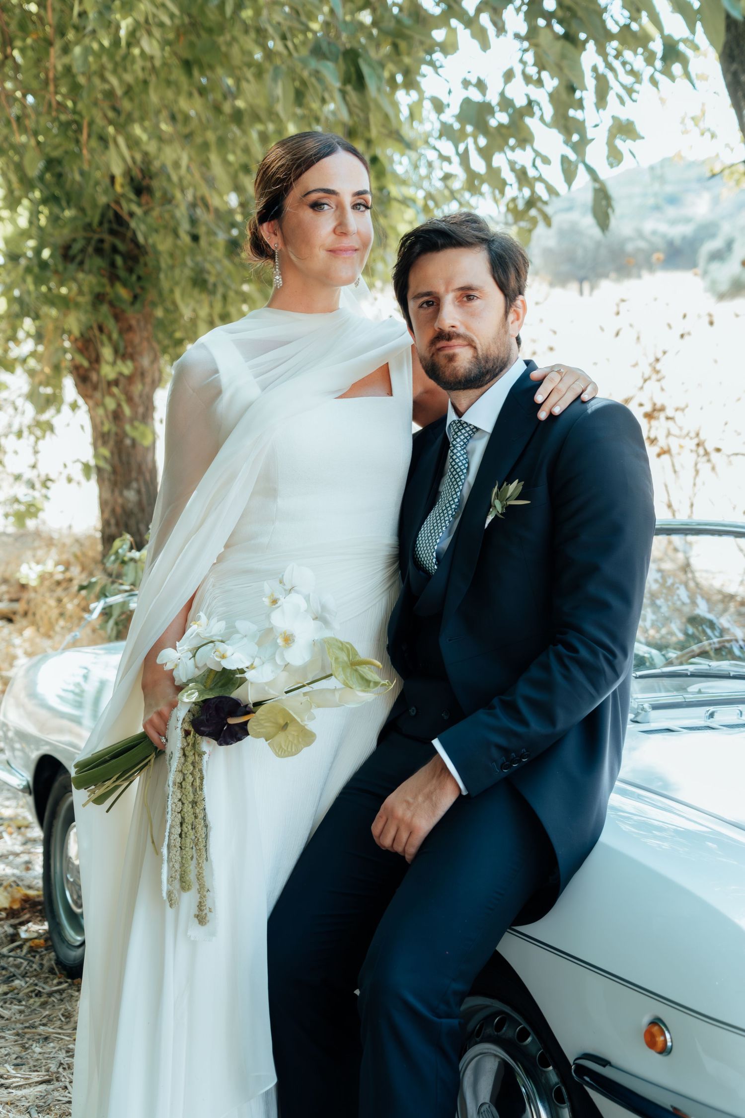 Elegant bride and groom posing beside a white vintage convertible car under a shady tree.
