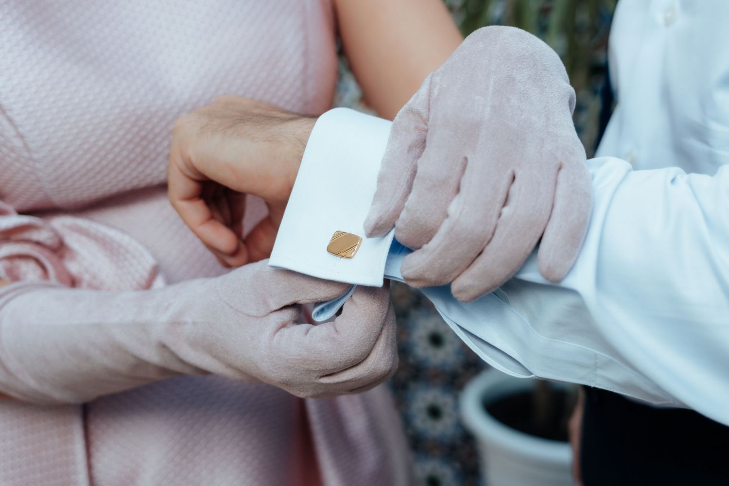 Close-up of woman fastening gold cufflink on groom's white dress shirt sleeve outdoors.