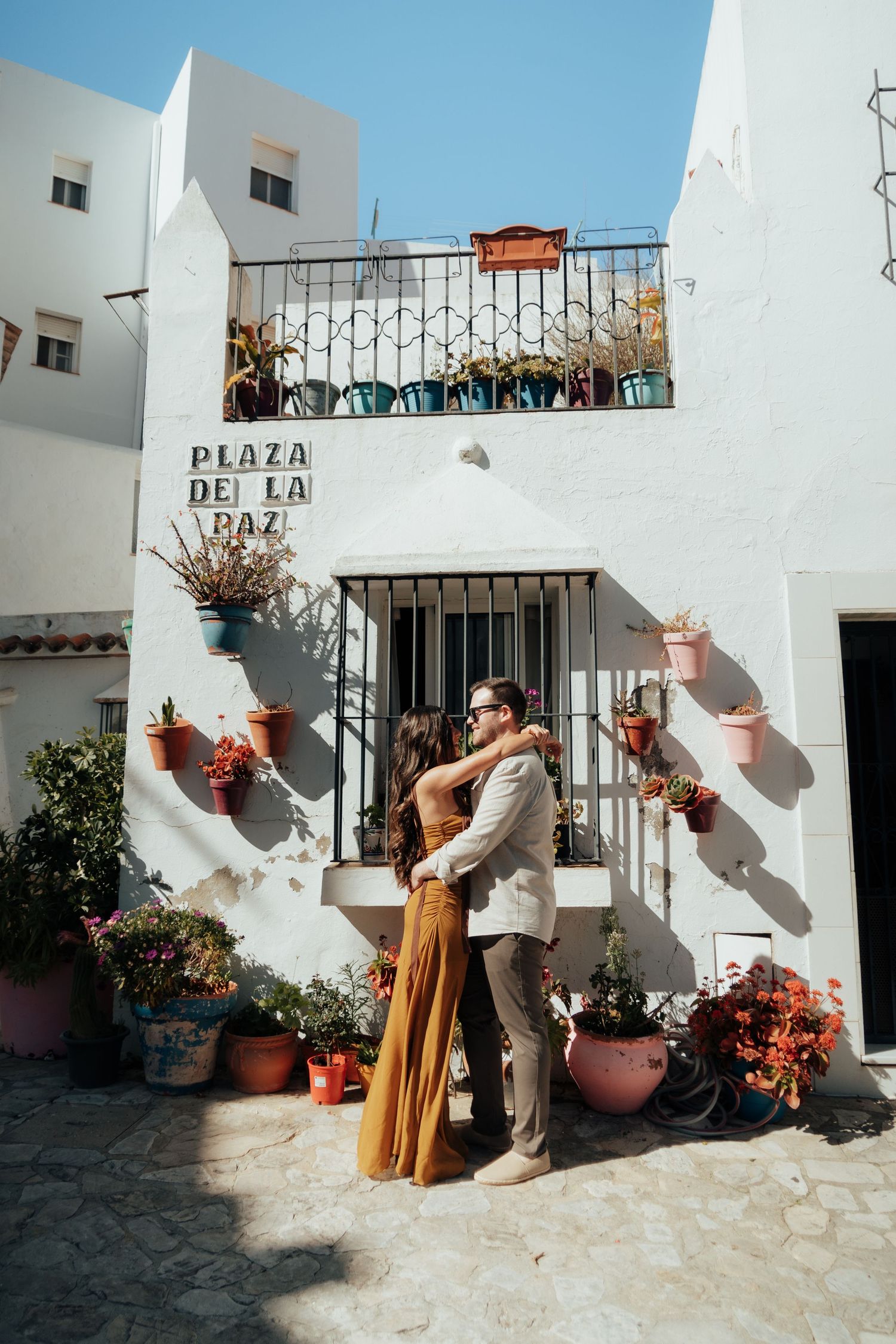 Couple embracing at Plaza de la Paz, Spain, surrounded by colorful potted plants on a white Mediterranean building.
