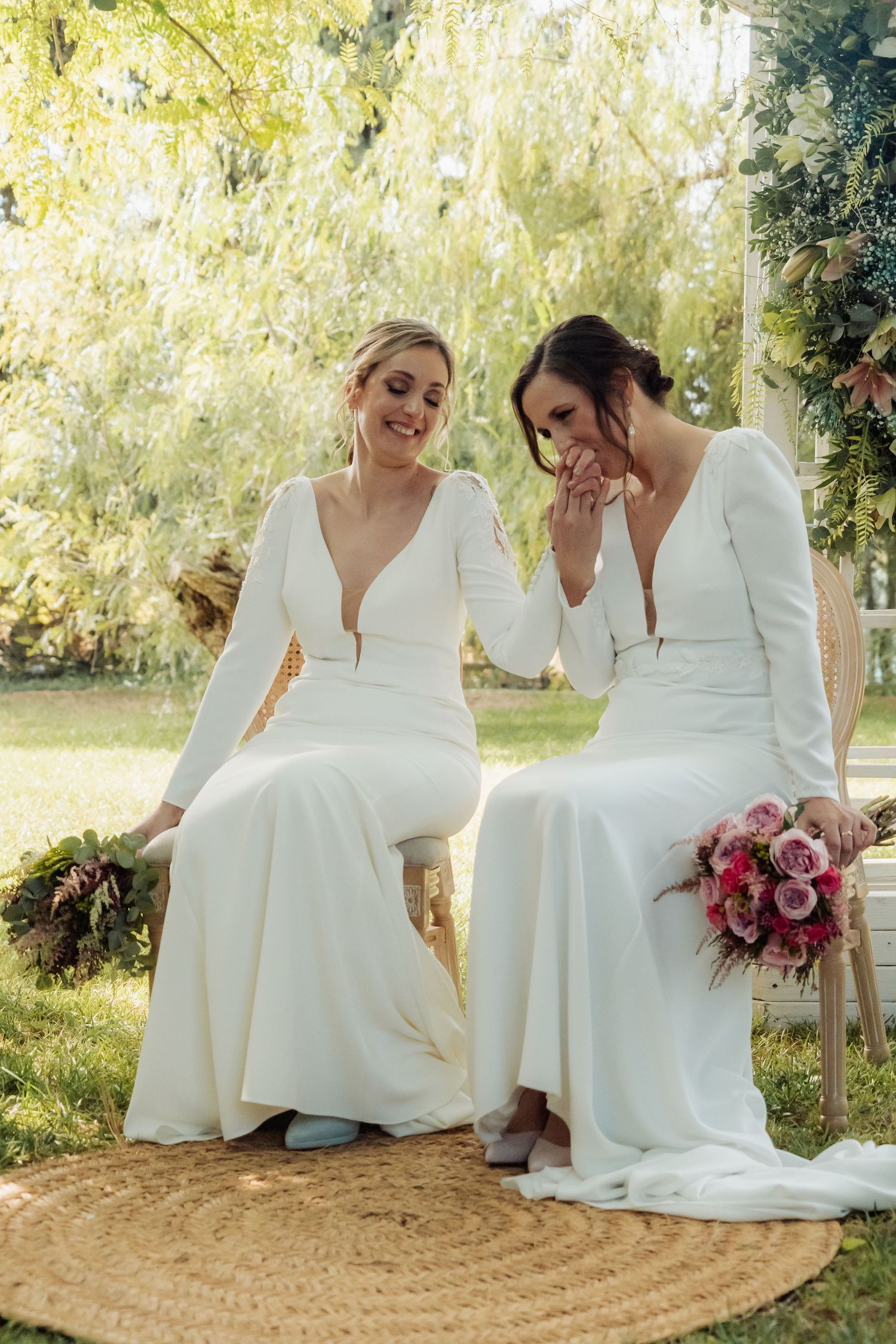 Two brides in elegant white dresses sharing a tender moment seated under a garden floral arch.