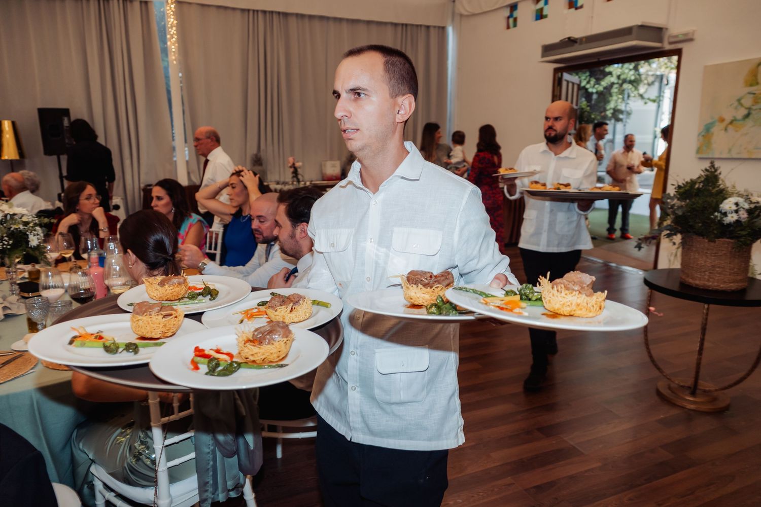 Waiter carrying multiple elegantly plated dishes to guests at a busy wedding reception venue.