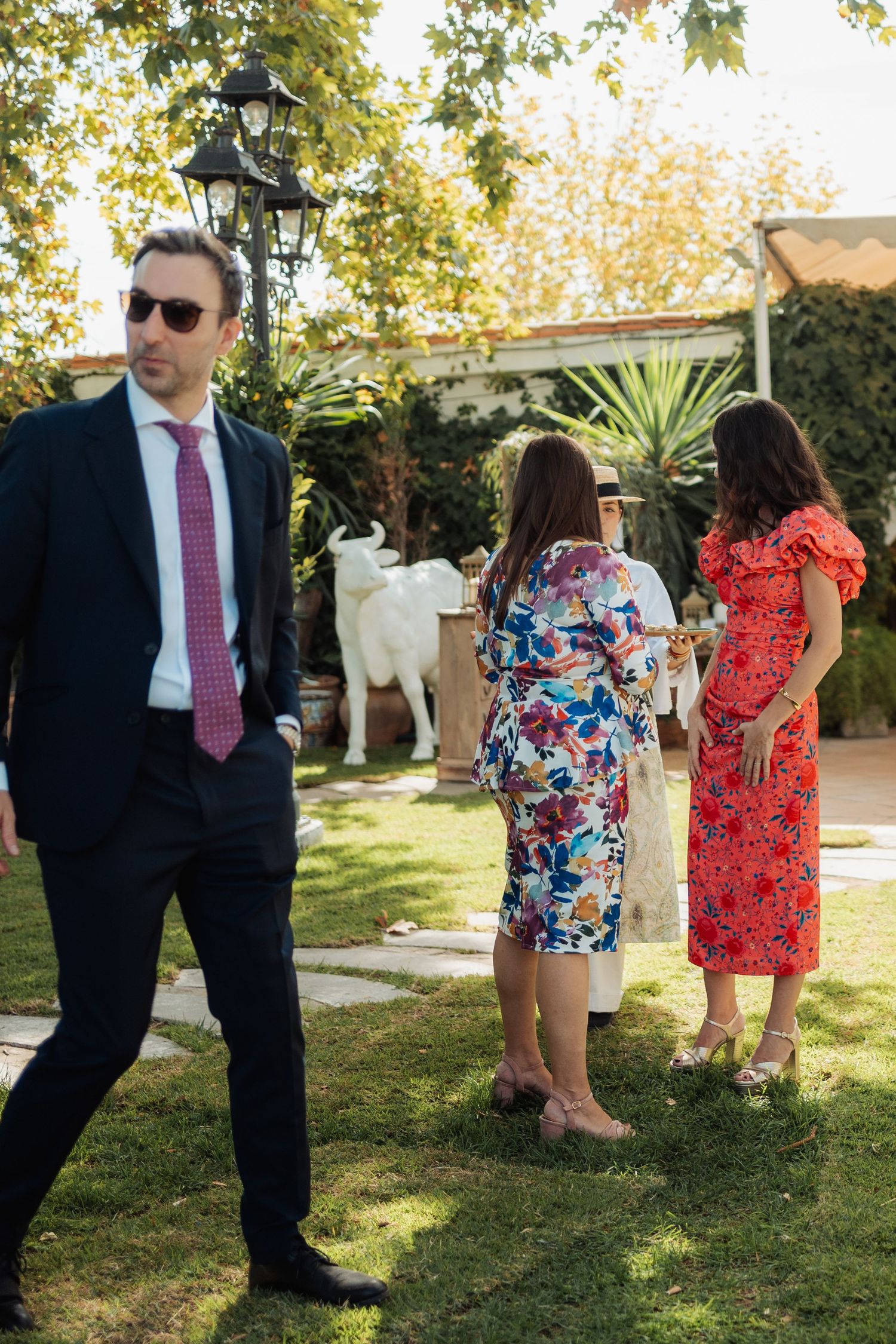 Wedding guests in colorful attire mingle in garden near white bull sculpture at venue.