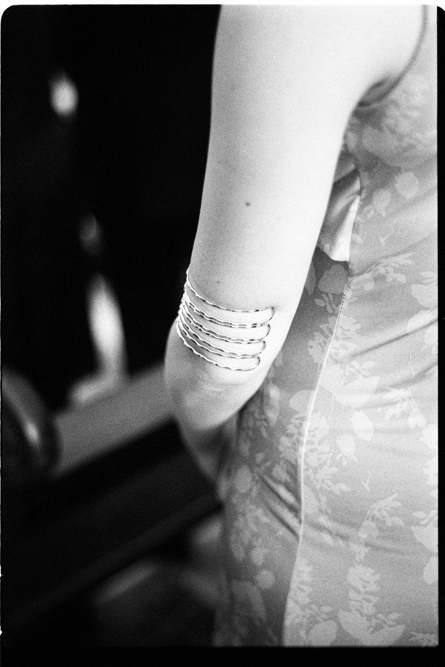 Black and white close-up of bridesmaid wearing multiple silver bangle bracelets on arm in floral dress.