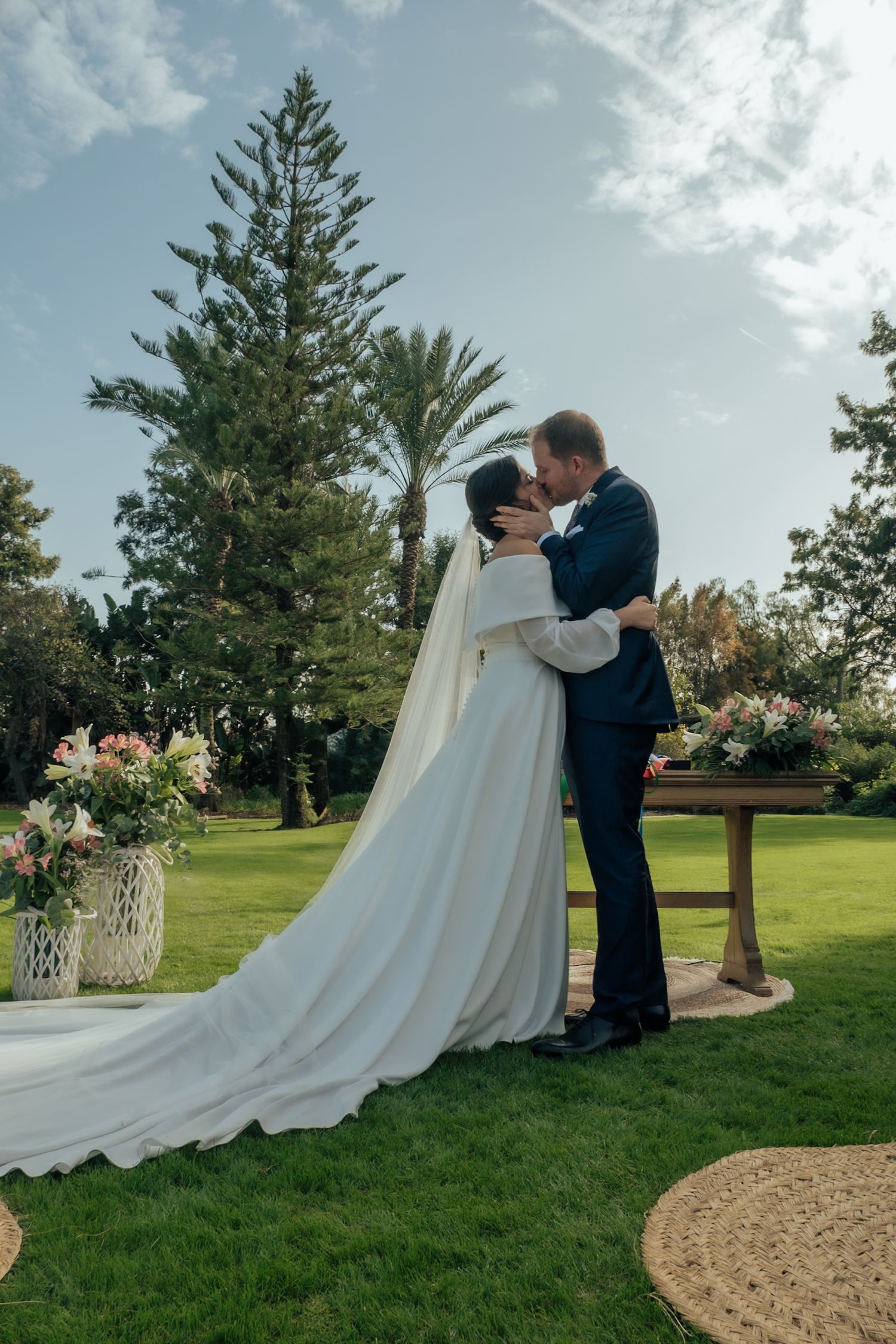 Bride and groom sharing their first kiss at an outdoor garden wedding ceremony with palm trees and flowers.