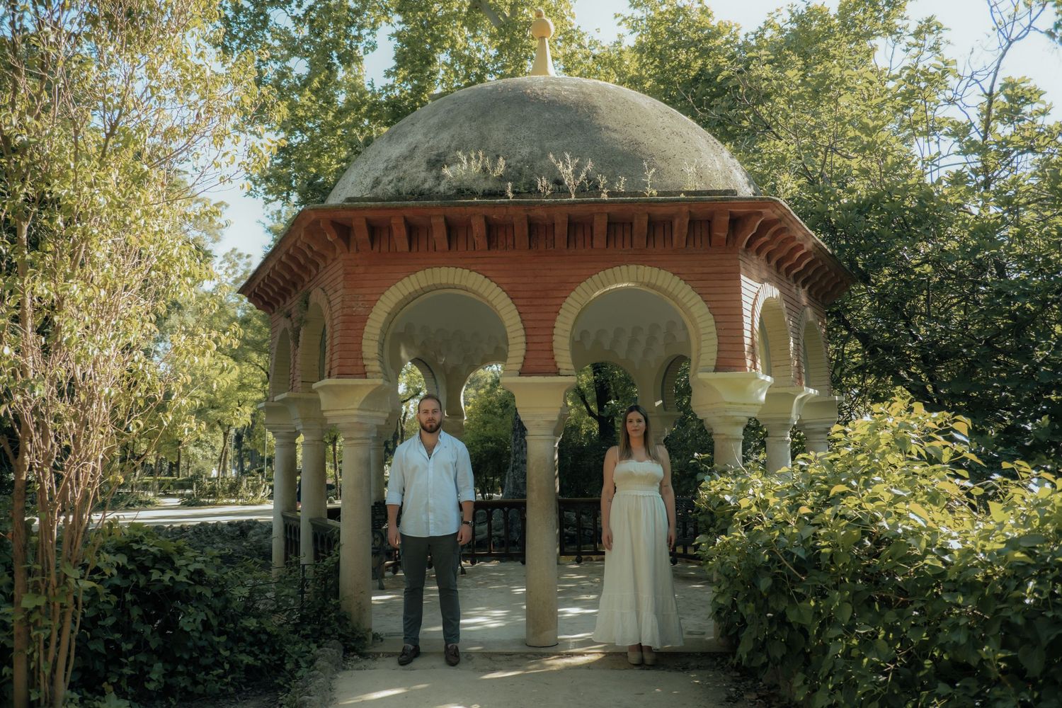Couple standing before ornate Moorish pavilion in lush Seville park garden, woman in white dress.