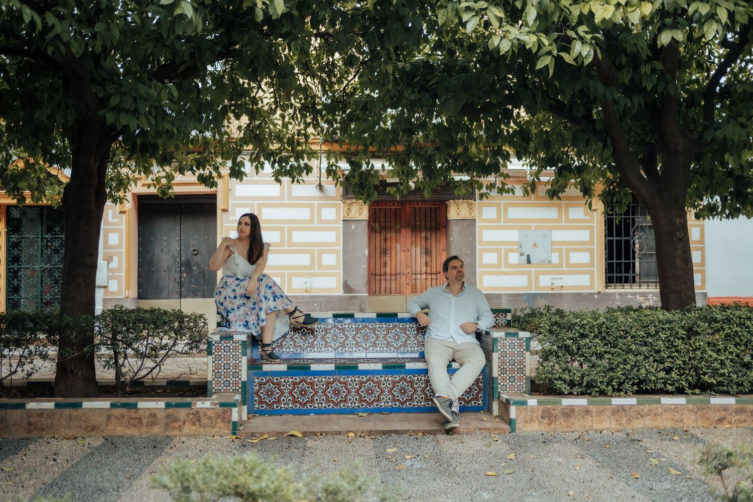 Couple sitting apart on a decorative tiled bench surrounded by trees outside a colorful building.
