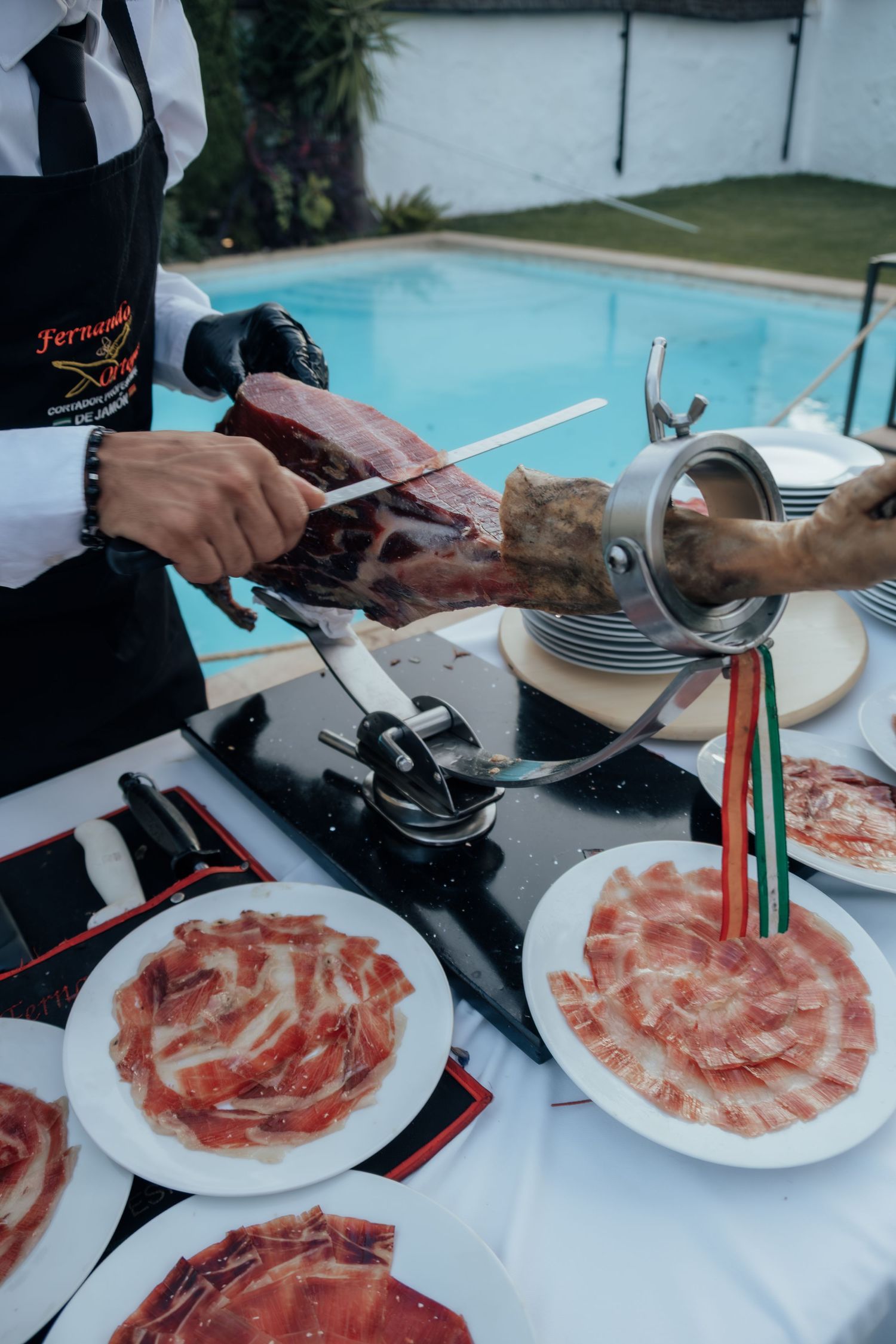 Catering staff slicing traditional Spanish jamon iberico ham beside a pool at wedding reception.