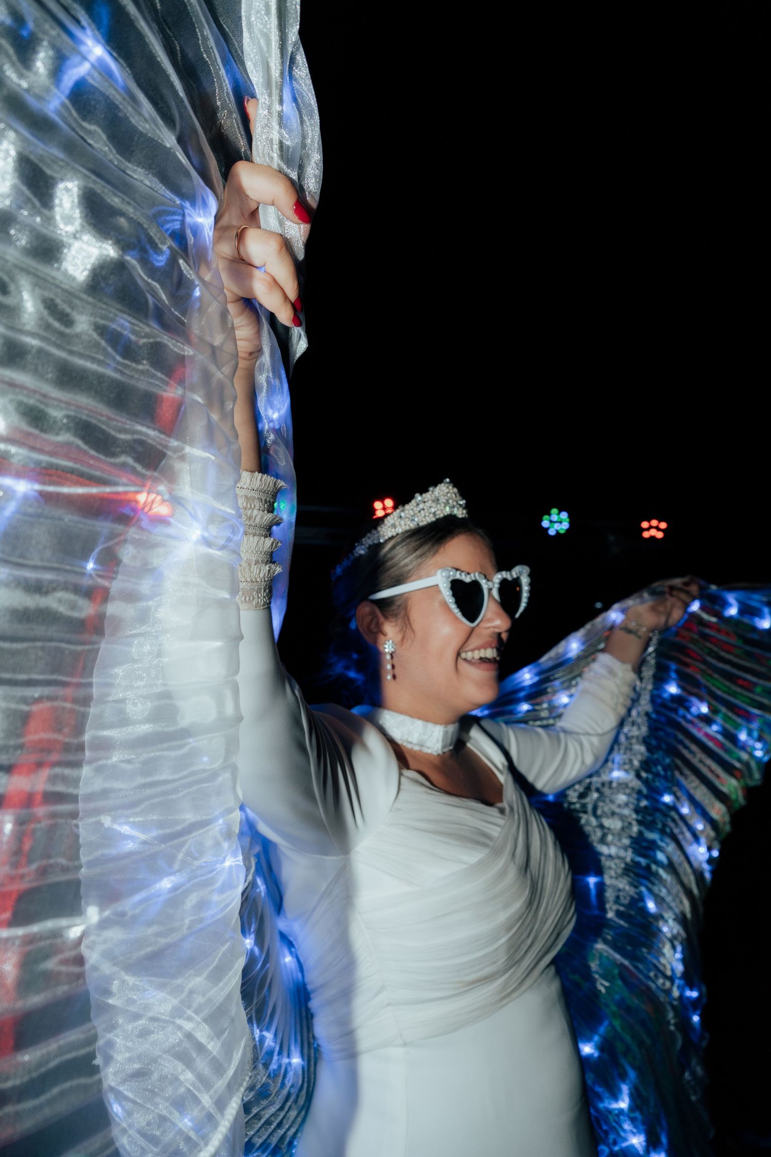 A bride in white wearing heart-shaped sunglasses and a tiara poses with illuminated LED wings at night.