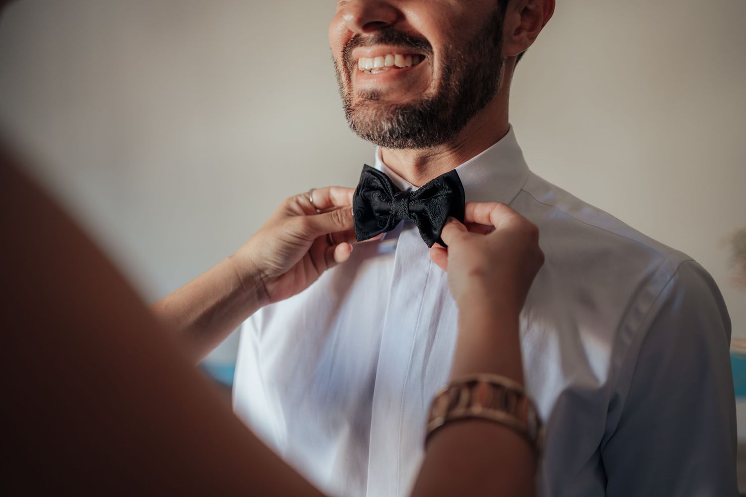 Close-up of a woman's hands adjusting a black bow tie on a smiling bearded groom in a white shirt.