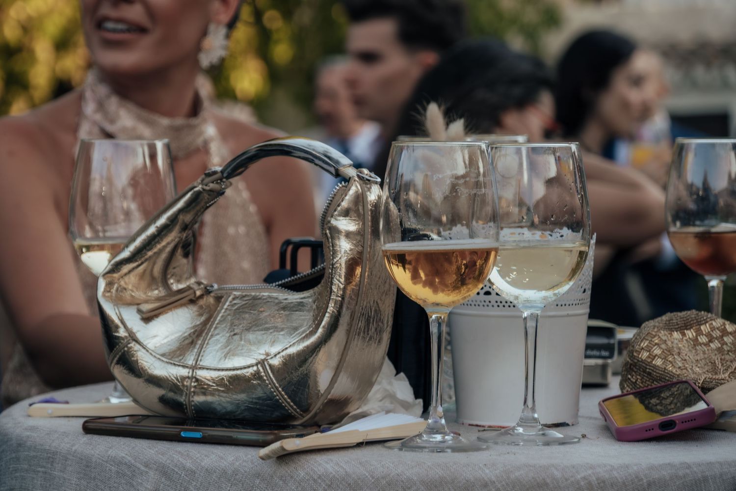 Silver designer handbag beside wine glasses on wedding reception table with guests in background.