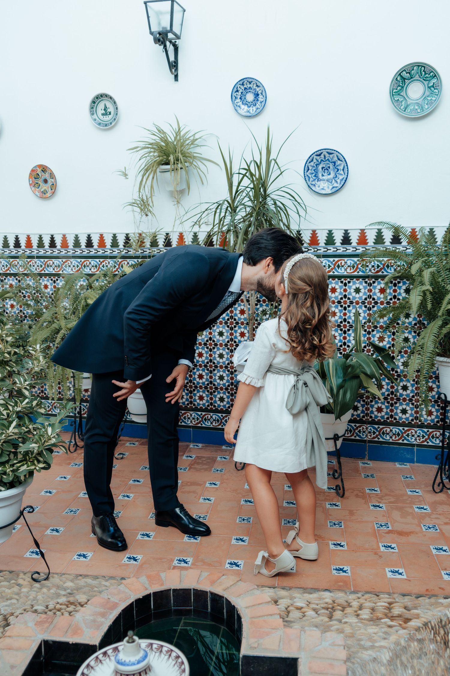 Groom in dark suit bending to kiss young flower girl in white dress in Spanish courtyard with decorative tiles.