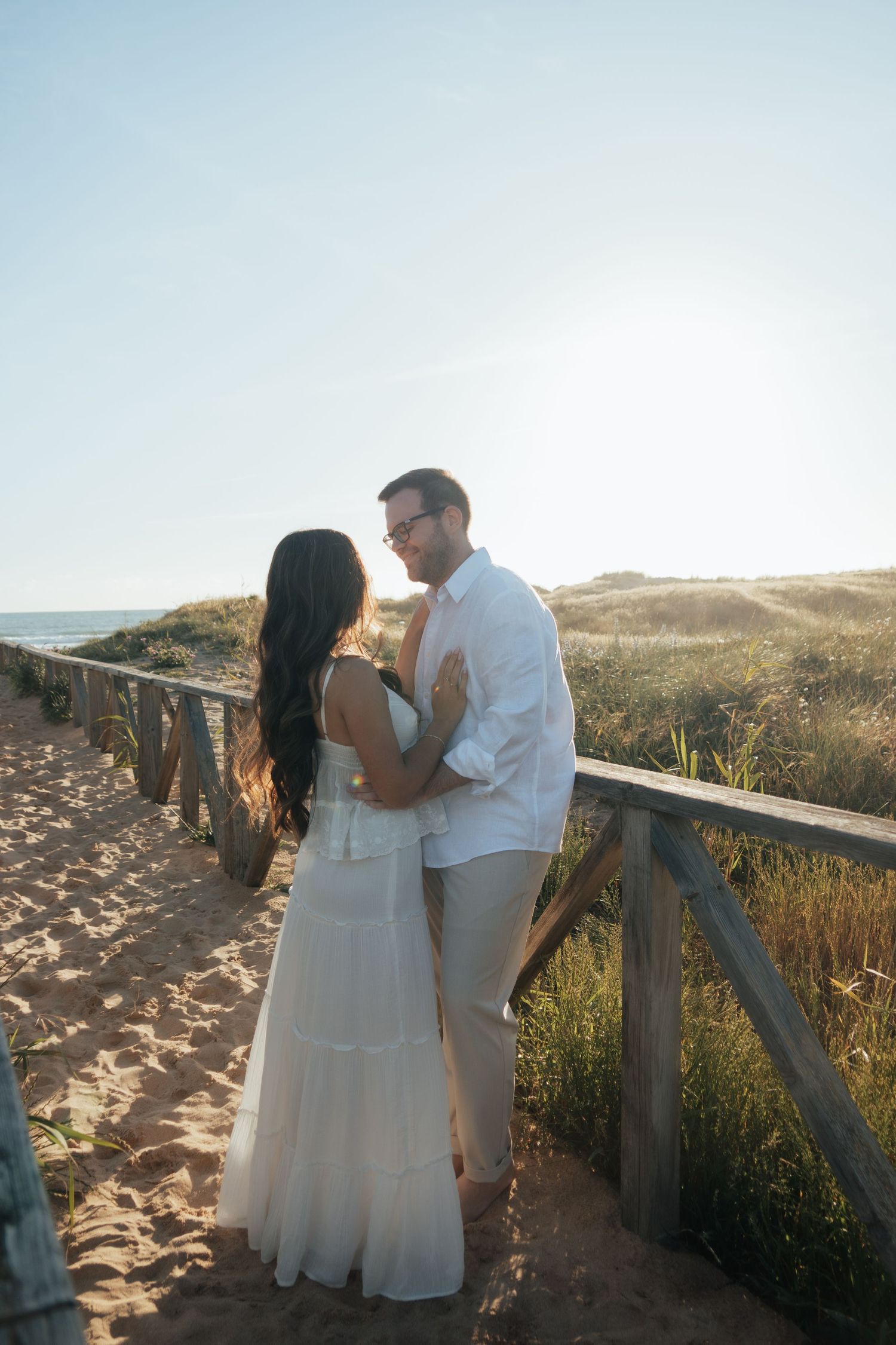 Couple in white outfits embracing on a sandy beach boardwalk at golden hour with ocean views.