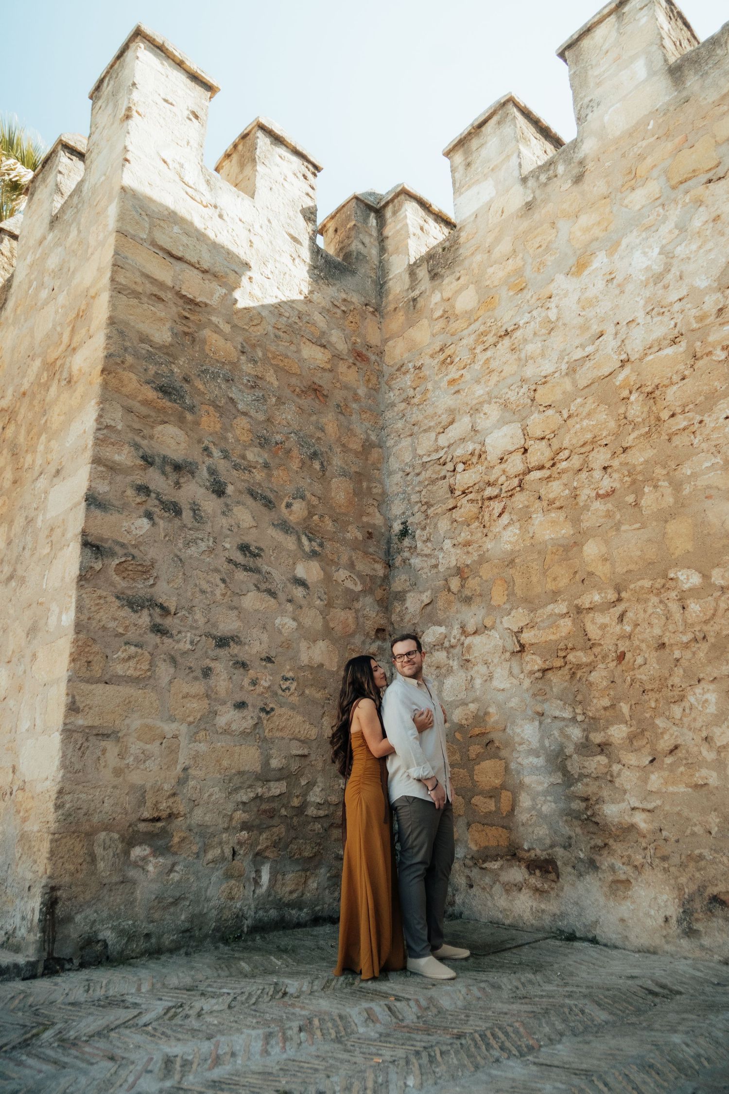 Couple holding hands standing against ancient stone castle walls with battlements in warm sunlight.