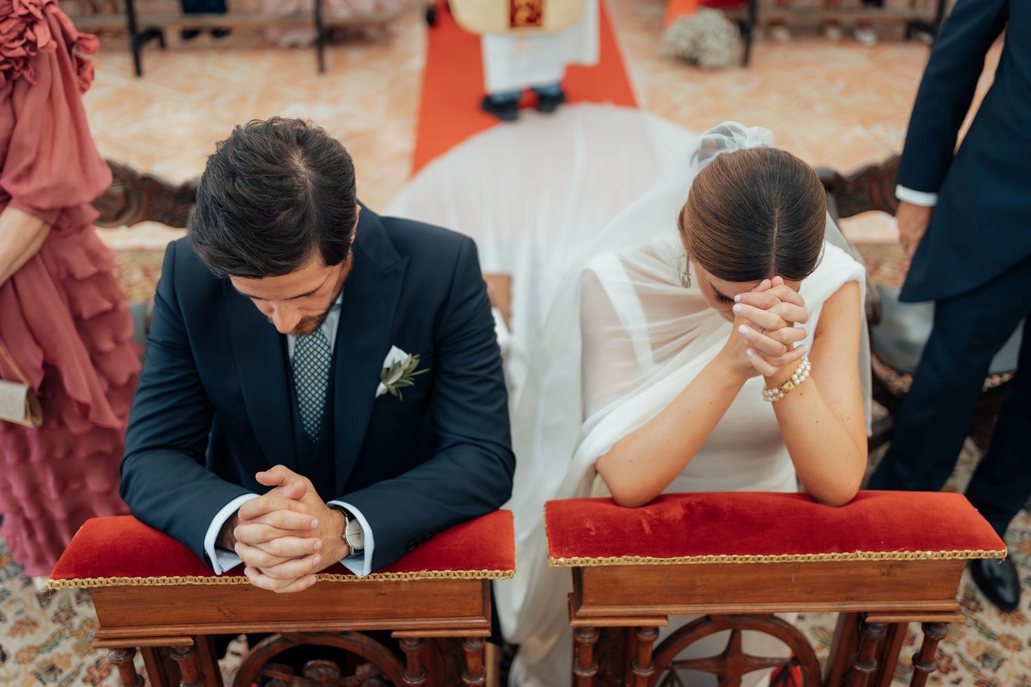Bride and groom kneeling in prayer at red kneelers during Catholic wedding ceremony in church.