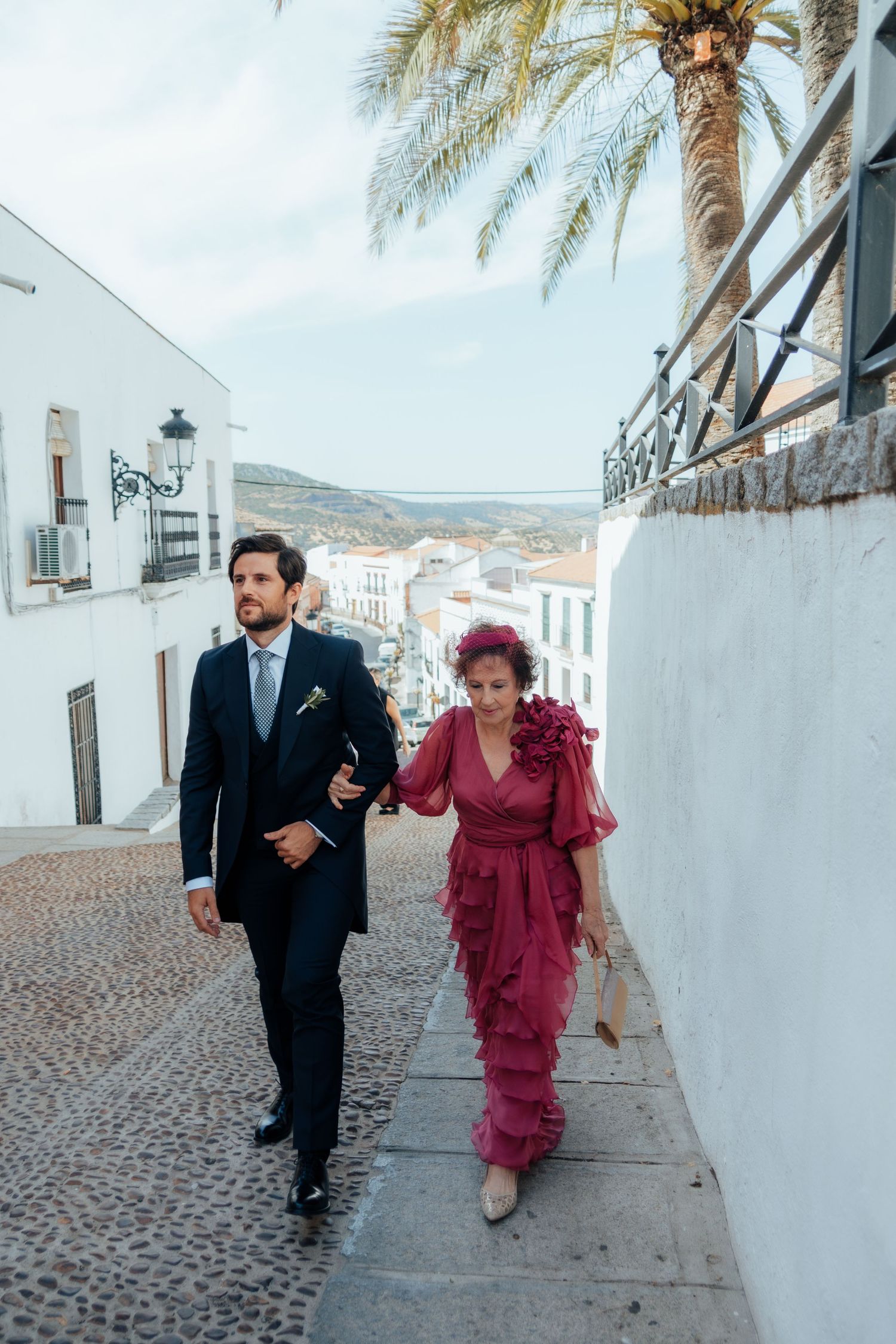 Groom in navy suit escorting woman in pink ruffled dress along cobblestone street in Spanish village.
