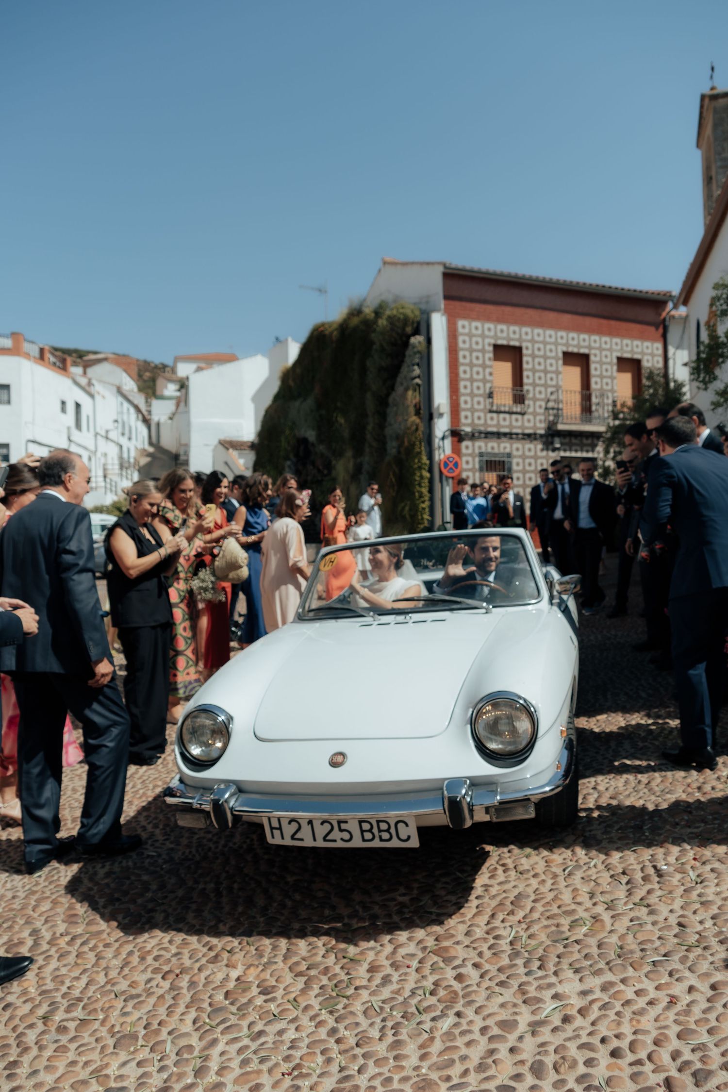 Bride and groom departing in a white vintage convertible car on cobblestone street surrounded by wedding guests.