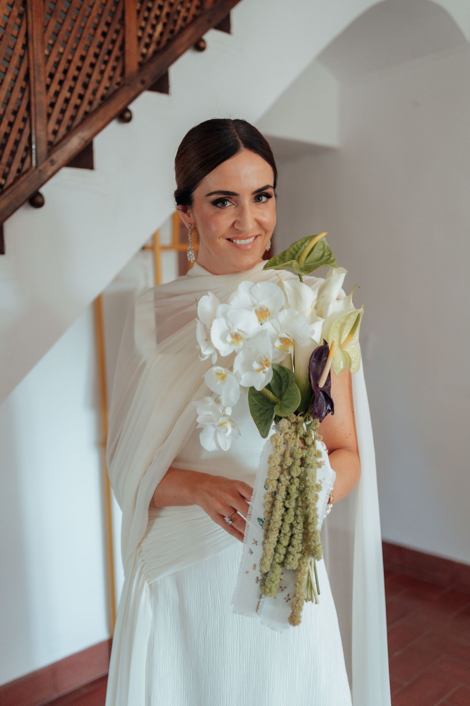 Smiling bride in white gown holding tropical orchid bouquet with dried hanging flowers on staircase.