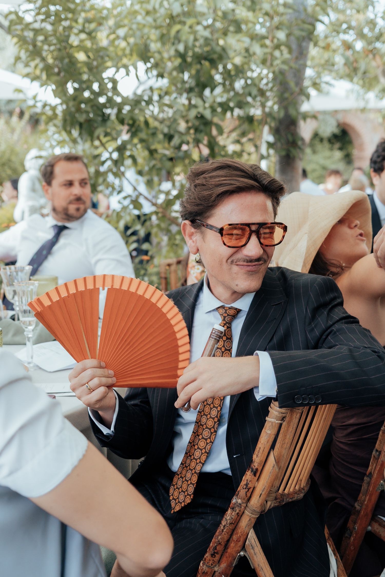 Stylish wedding guest in pinstripe suit and sunglasses holding orange hand fan at outdoor reception.