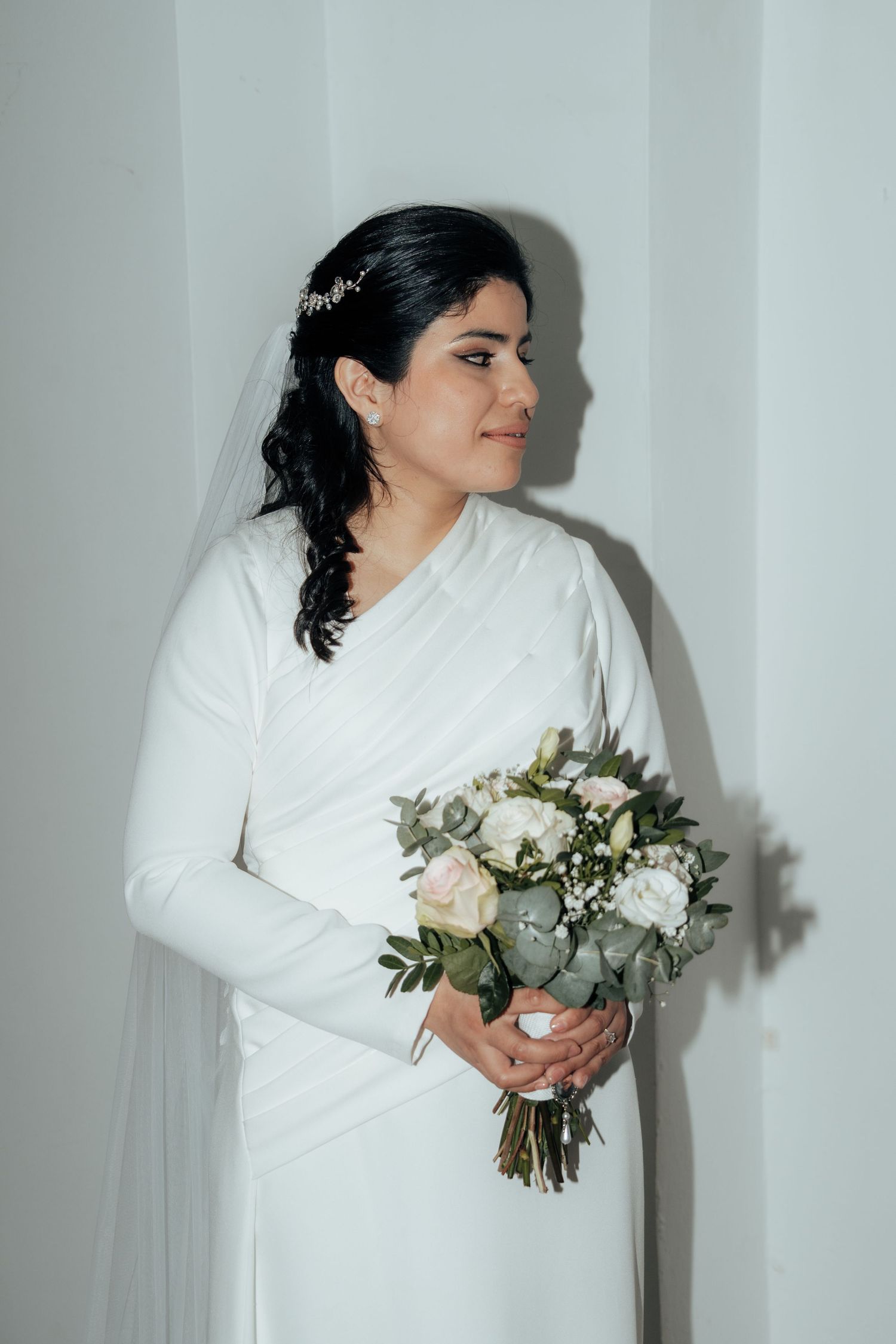 Elegant dark-haired bride in long white dress holding rose bouquet with crystal hair vine and veil.