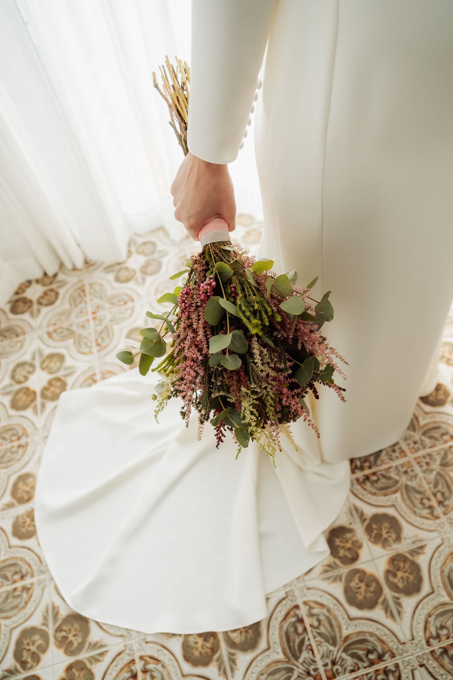 Bride in white gown holding rustic bouquet of eucalyptus and pink wildflowers by window.
