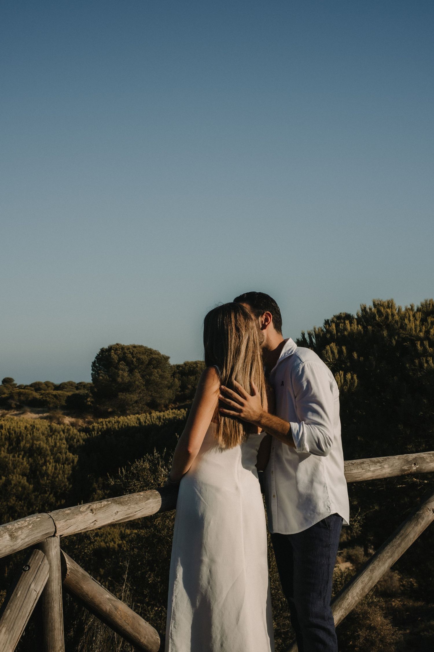 Couple sharing a tender kiss by a rustic wooden fence at golden hour in a scenic landscape.