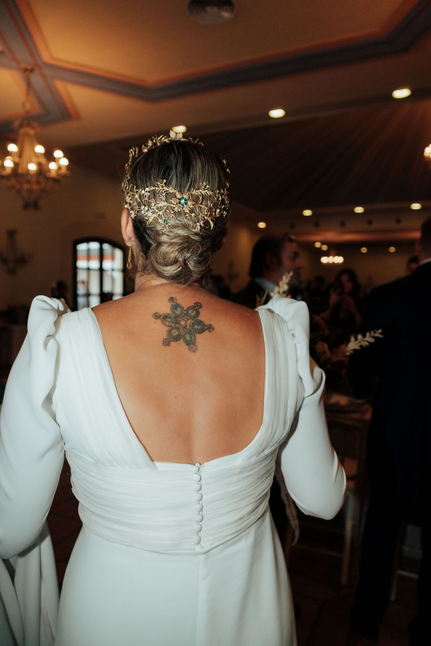 Bride in white backless wedding gown with floral crown and snowflake tattoo on back at indoor reception.