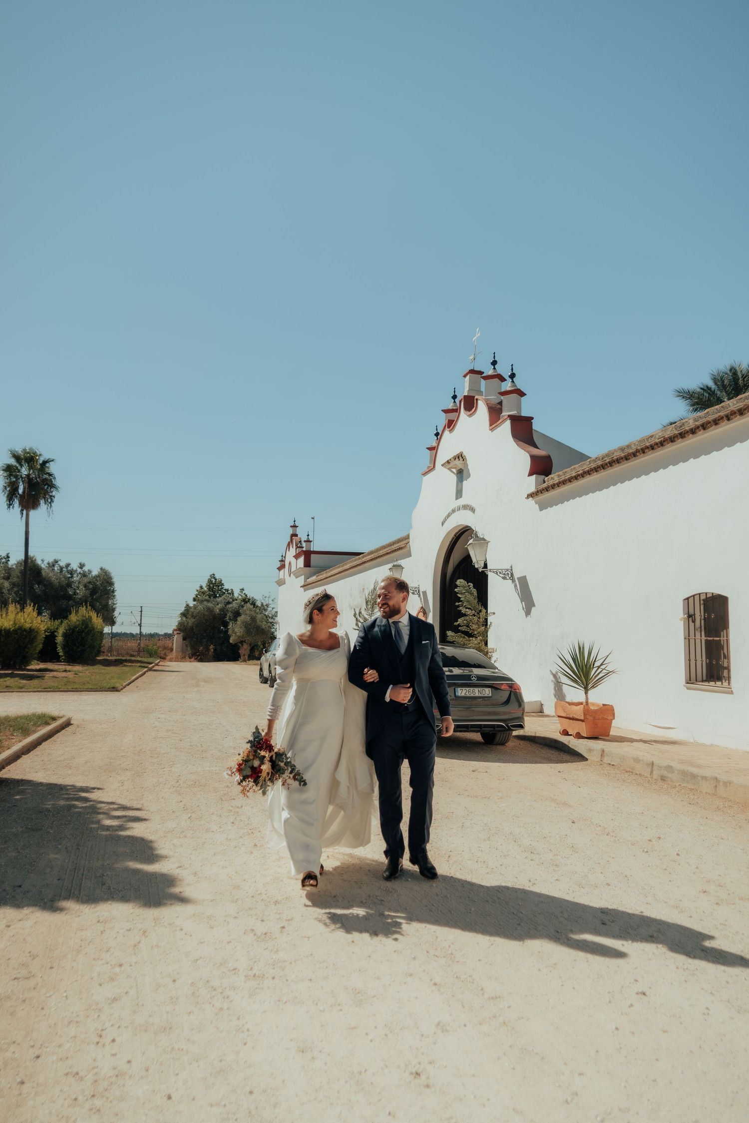 Newlyweds walk hand in arm along sandy path outside white Andalusian wedding venue under blue sky.