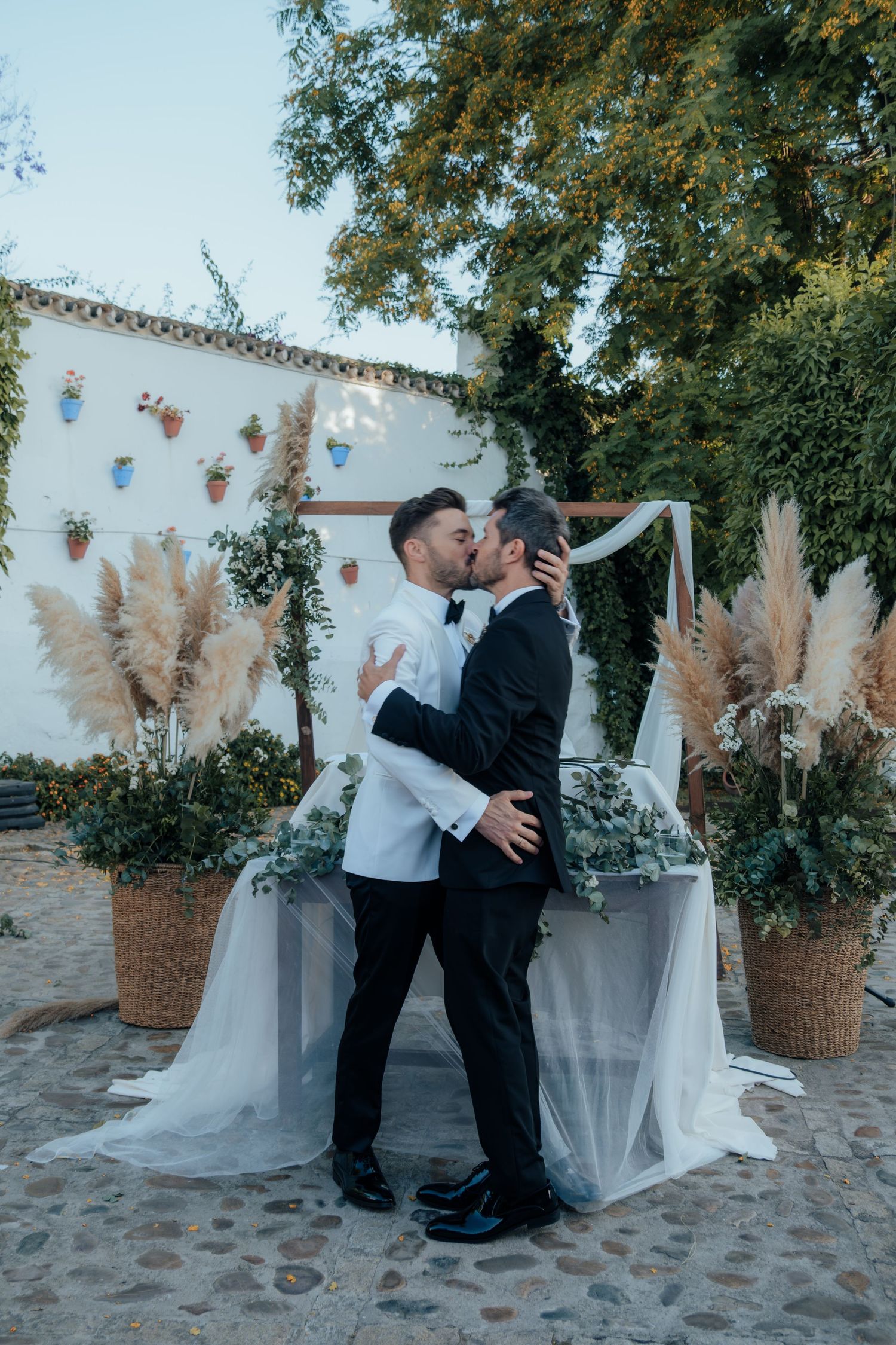 Two grooms share first kiss at altar decorated with pampas grass and greenery at outdoor wedding.