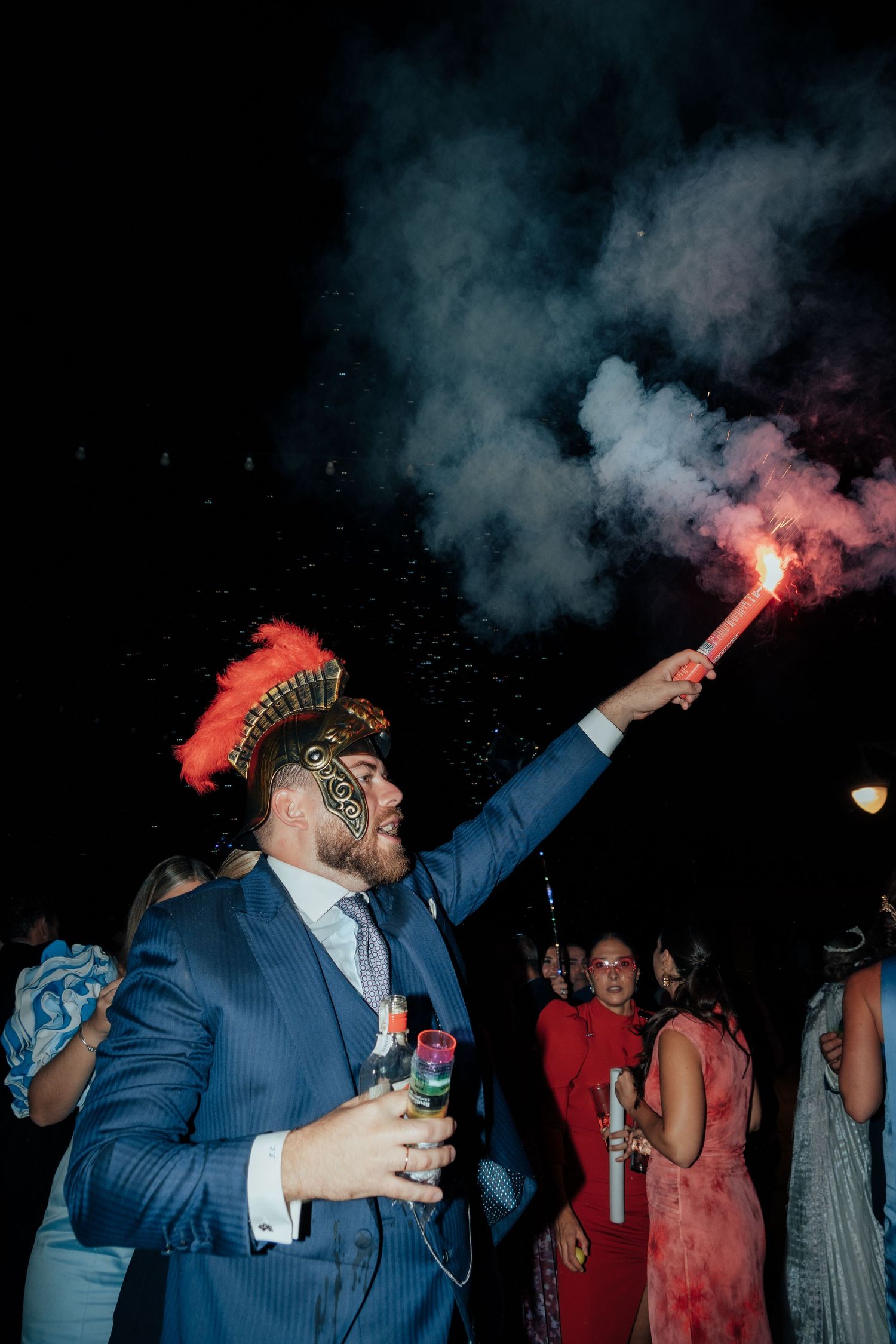 Wedding guest in blue suit holds a red flare and wears a decorative hat at a night celebration.