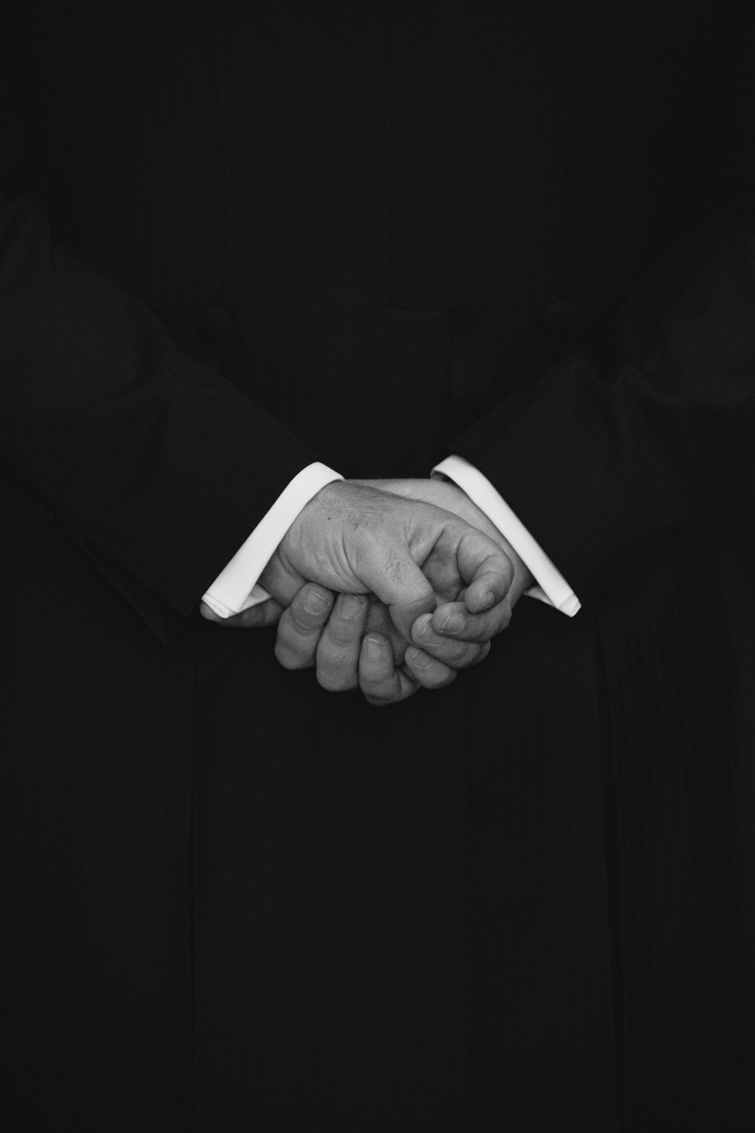 Black and white close-up of groom clasping hands behind back in formal suit.