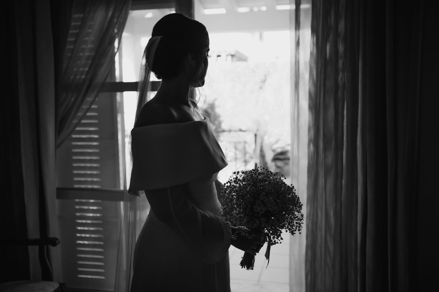 Black and white silhouette of a bride in an off-shoulder dress holding a bouquet while standing by a sunlit window.