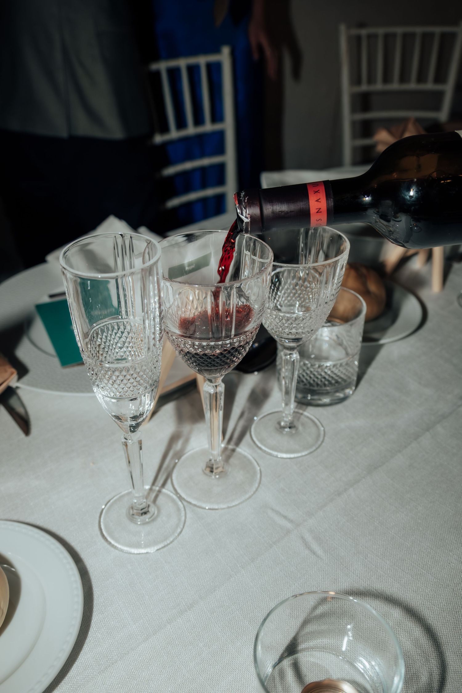Red wine being poured into crystal glass at elegant white linen wedding reception table setting.