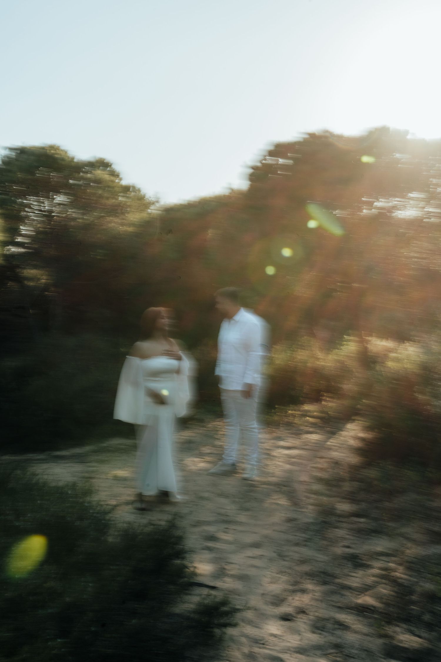 Blurry couple in white outfits standing on a sunlit path surrounded by trees with golden lens flare.