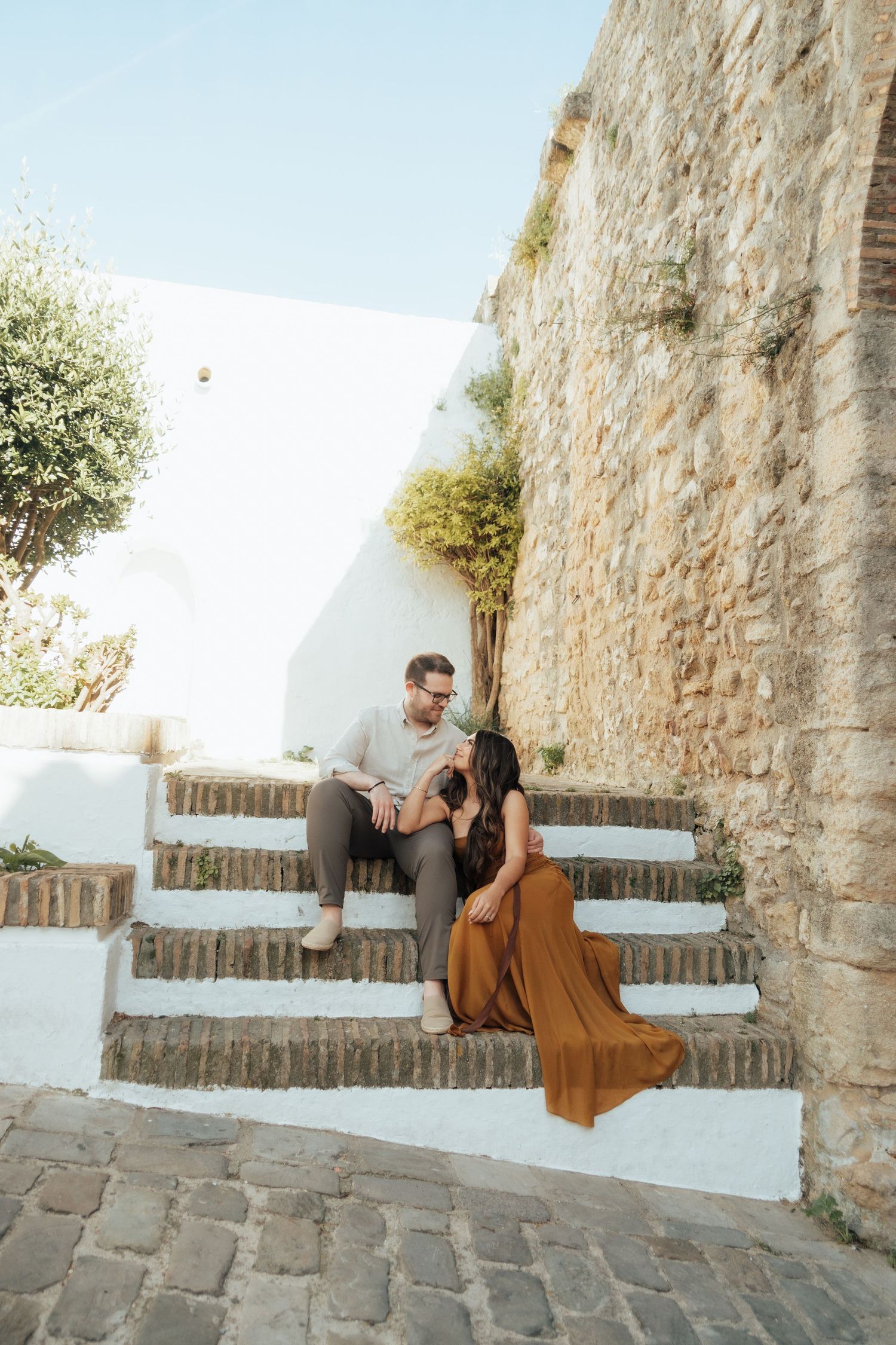 Couple sitting on rustic stone steps beside an ancient wall in a Mediterranean village setting.