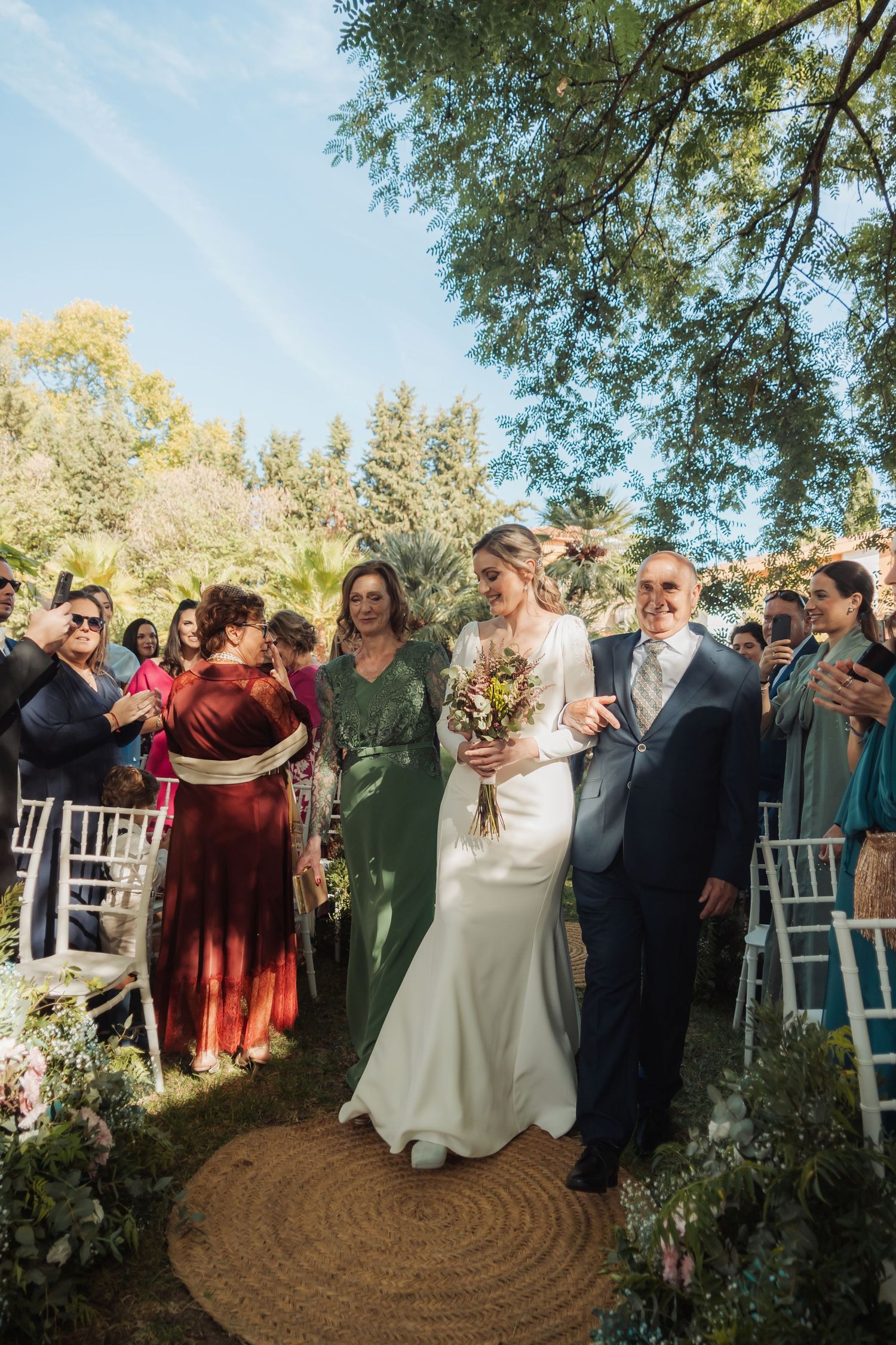 Smiling bride walks down the outdoor aisle escorted by parents as guests applaud and celebrate.