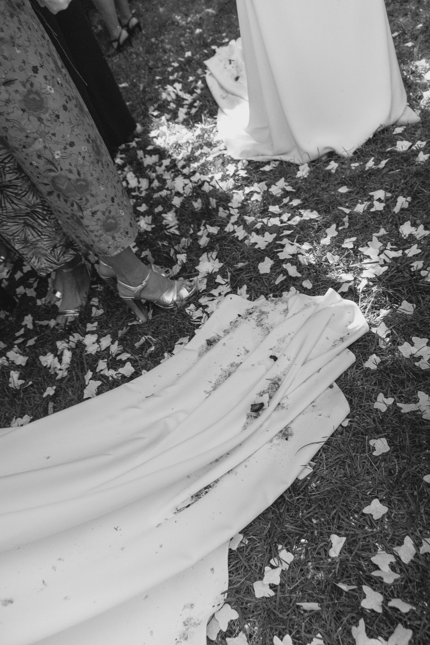 Black and white photo of bridal gown train lying on grass covered with confetti at outdoor wedding.