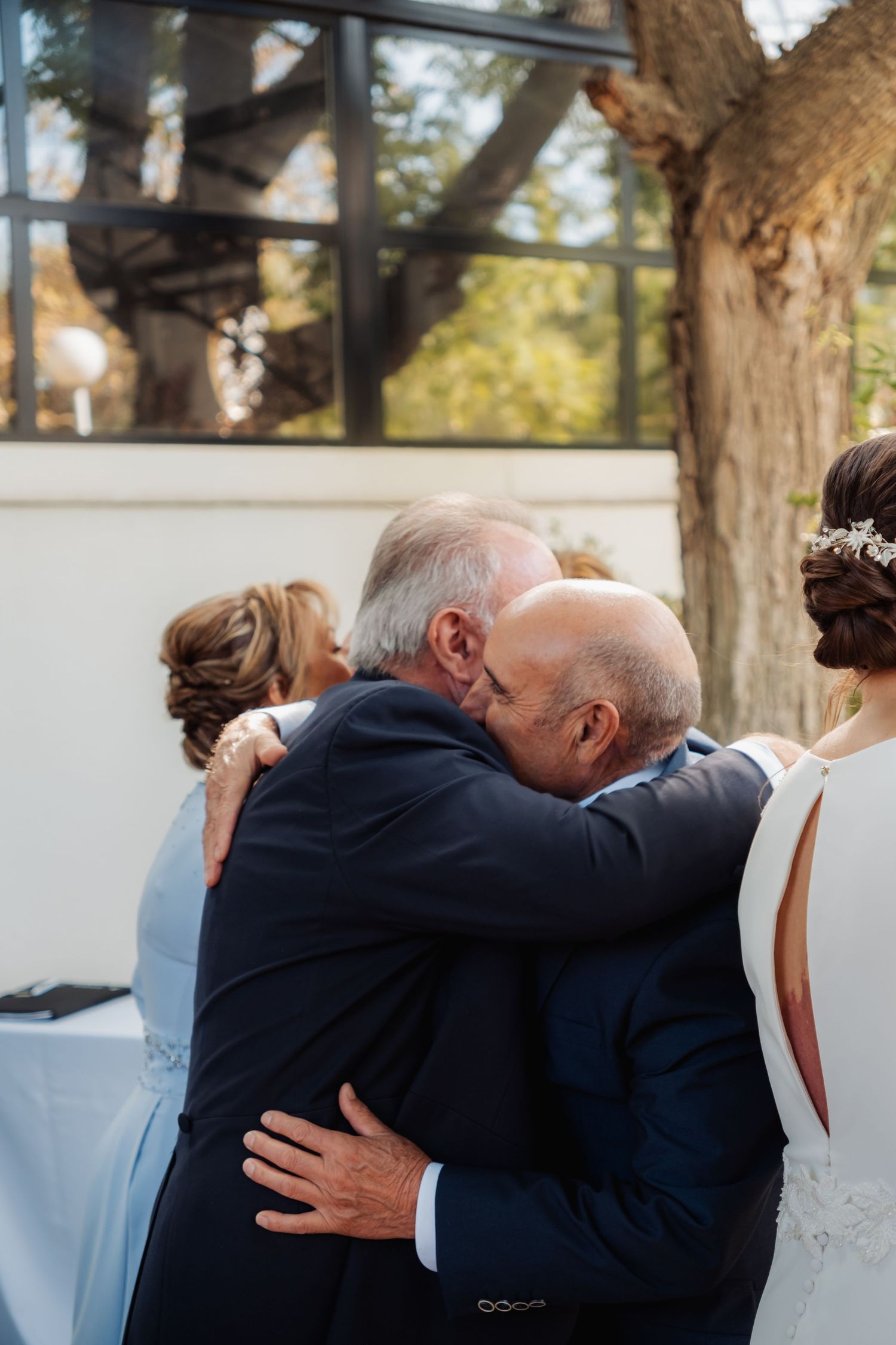 Two elderly men in navy suits share a heartfelt embrace at an outdoor wedding ceremony.