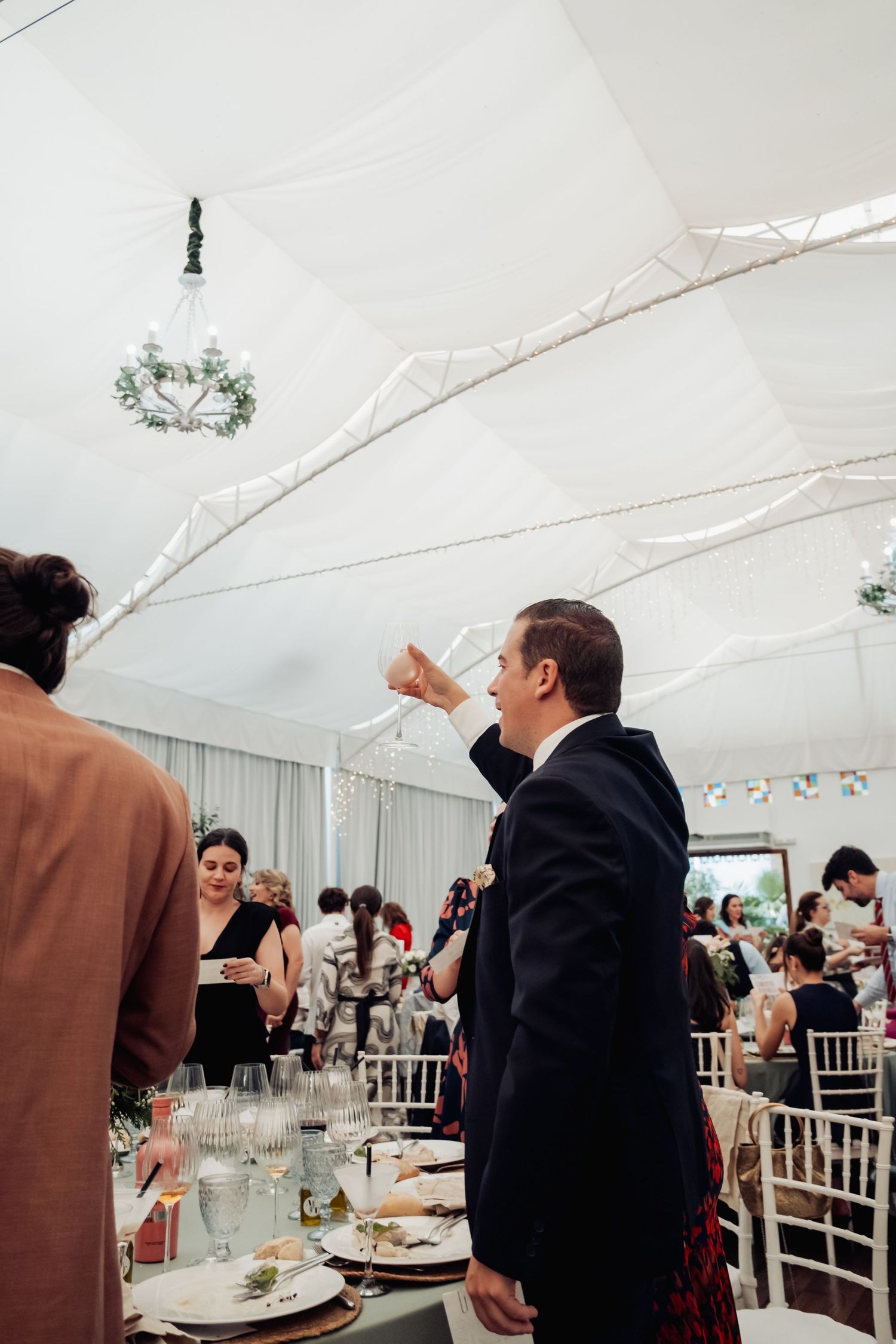 Man in dark suit raises glass for a toast at a decorated wedding reception under white tent.