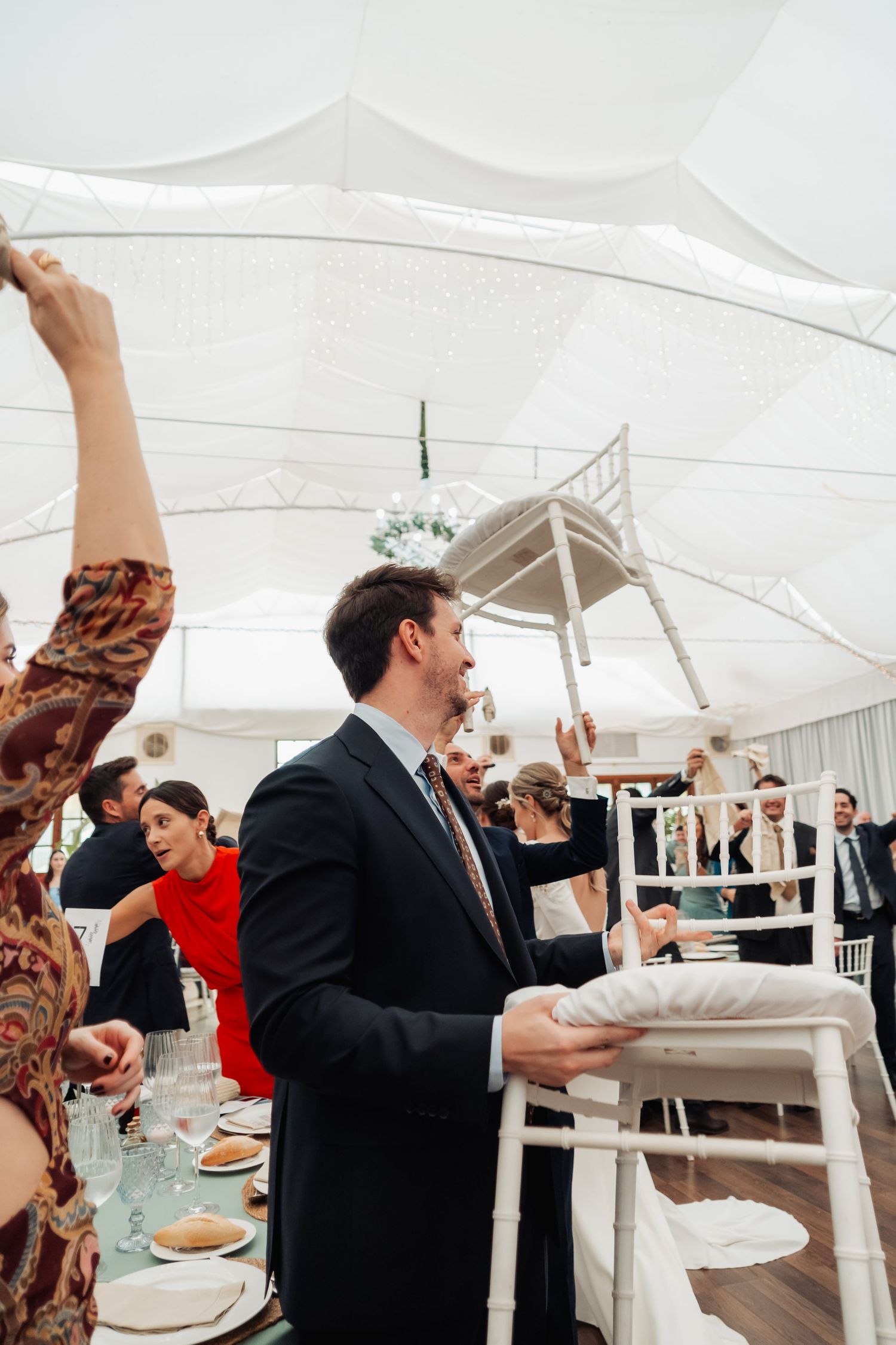 Wedding guests raising white chairs in joyful celebration inside a white draped marquee reception tent.