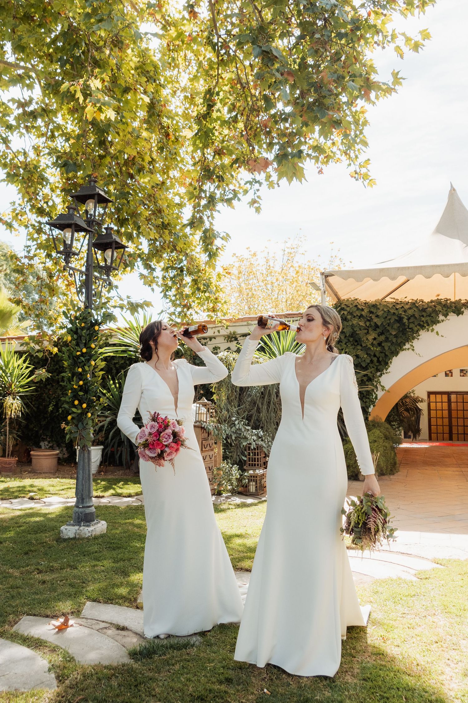 Two brides in elegant white long-sleeve gowns holding bouquets and drinks in a sunny garden.