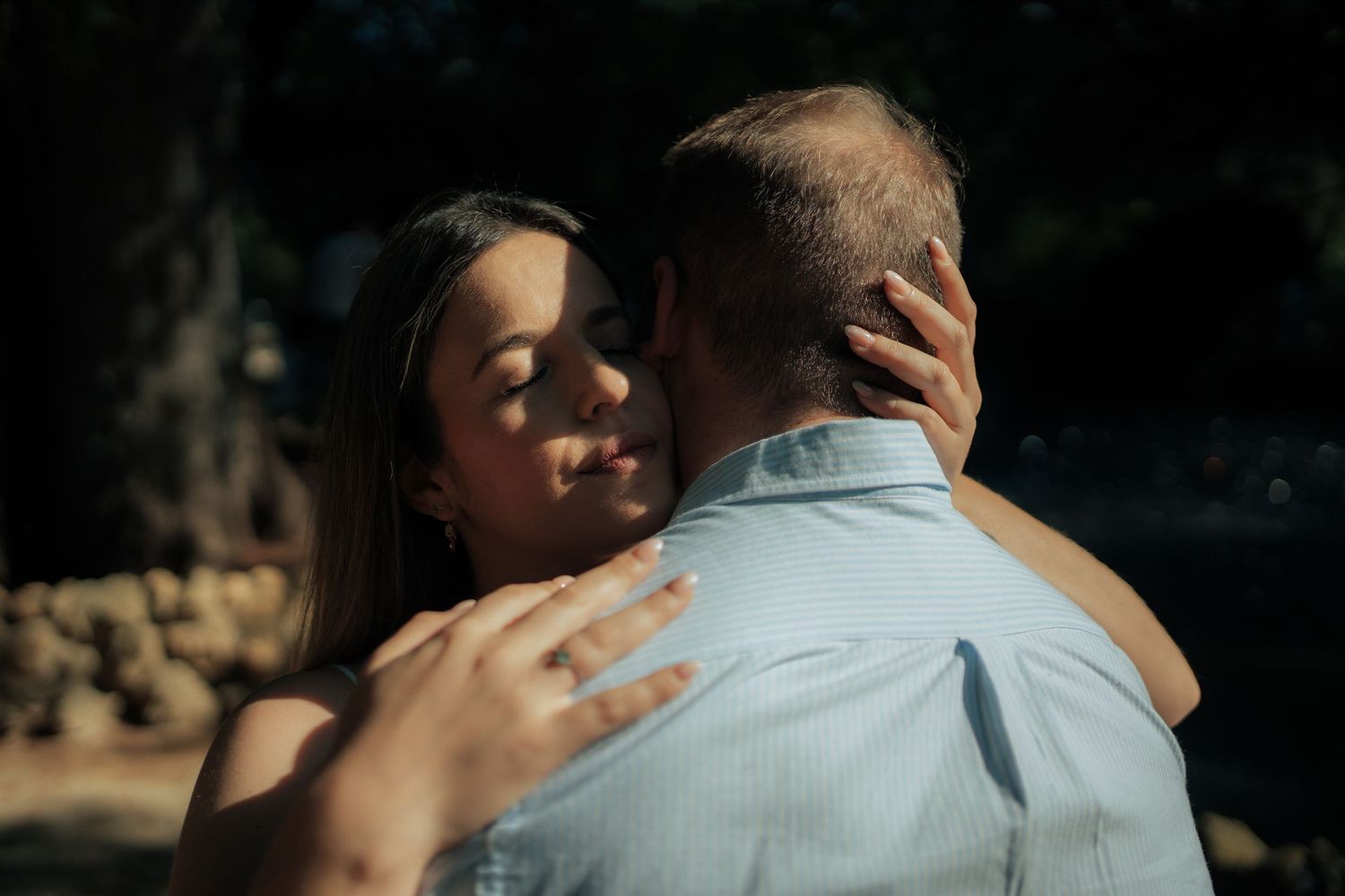 Close-up of woman with engagement ring embracing man in blue shirt in dappled sunlight.